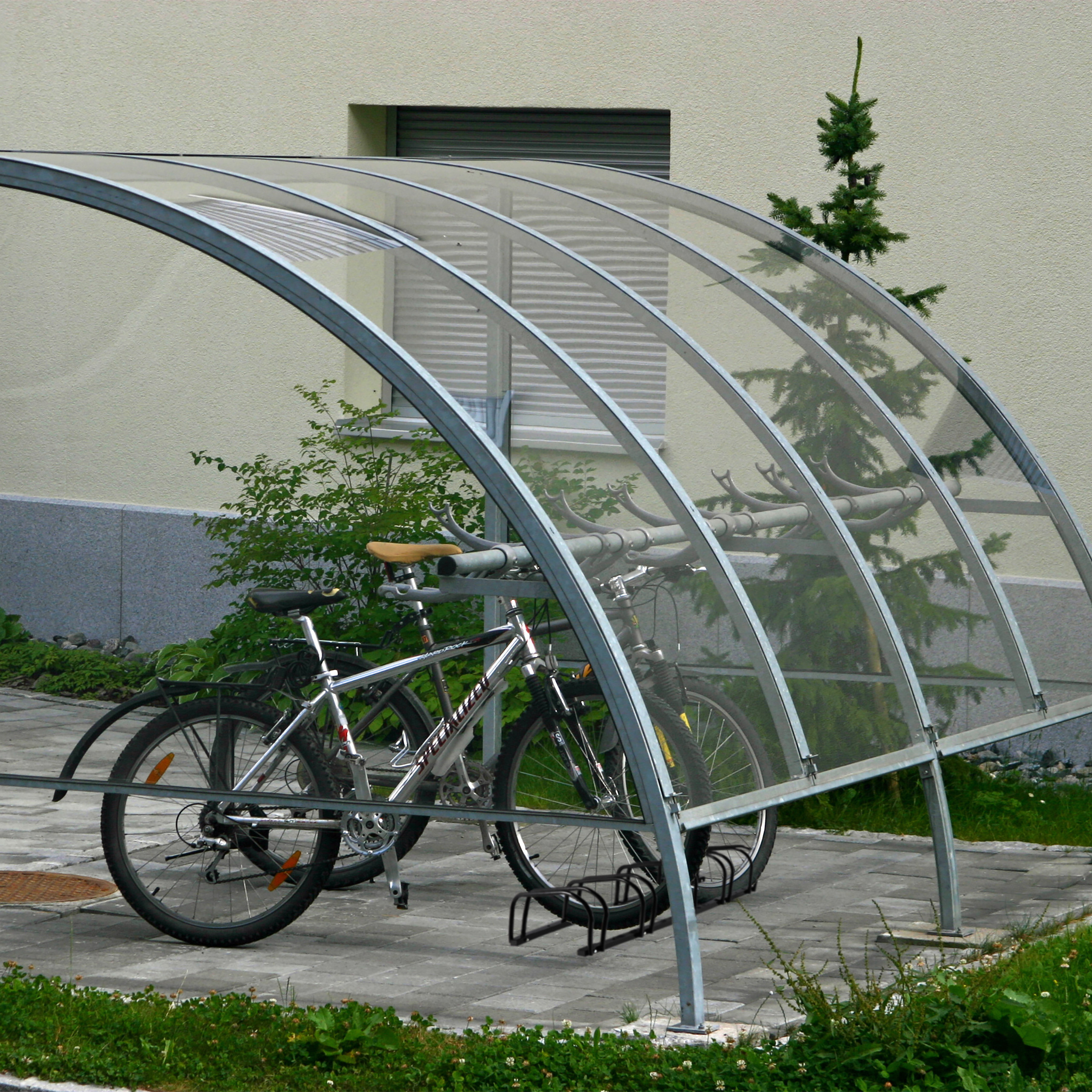 Zwei Fahrräder unter einem gewölbten Glasunterstand neben einem hellen Gebäude.