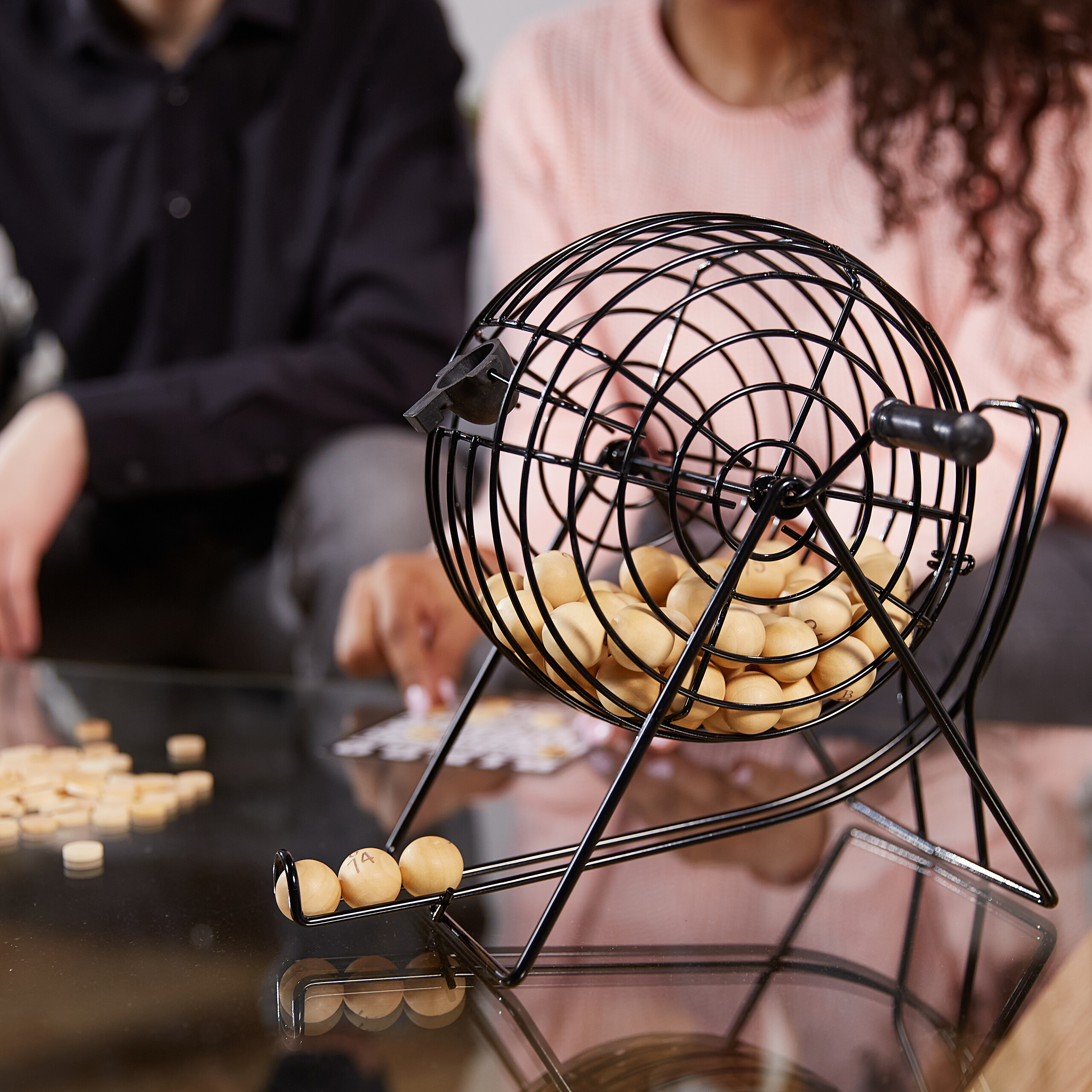 Eine schwarze Tombola-Trommel mit Holzkugeln auf einem Glastisch, dahinter zwei Personen.