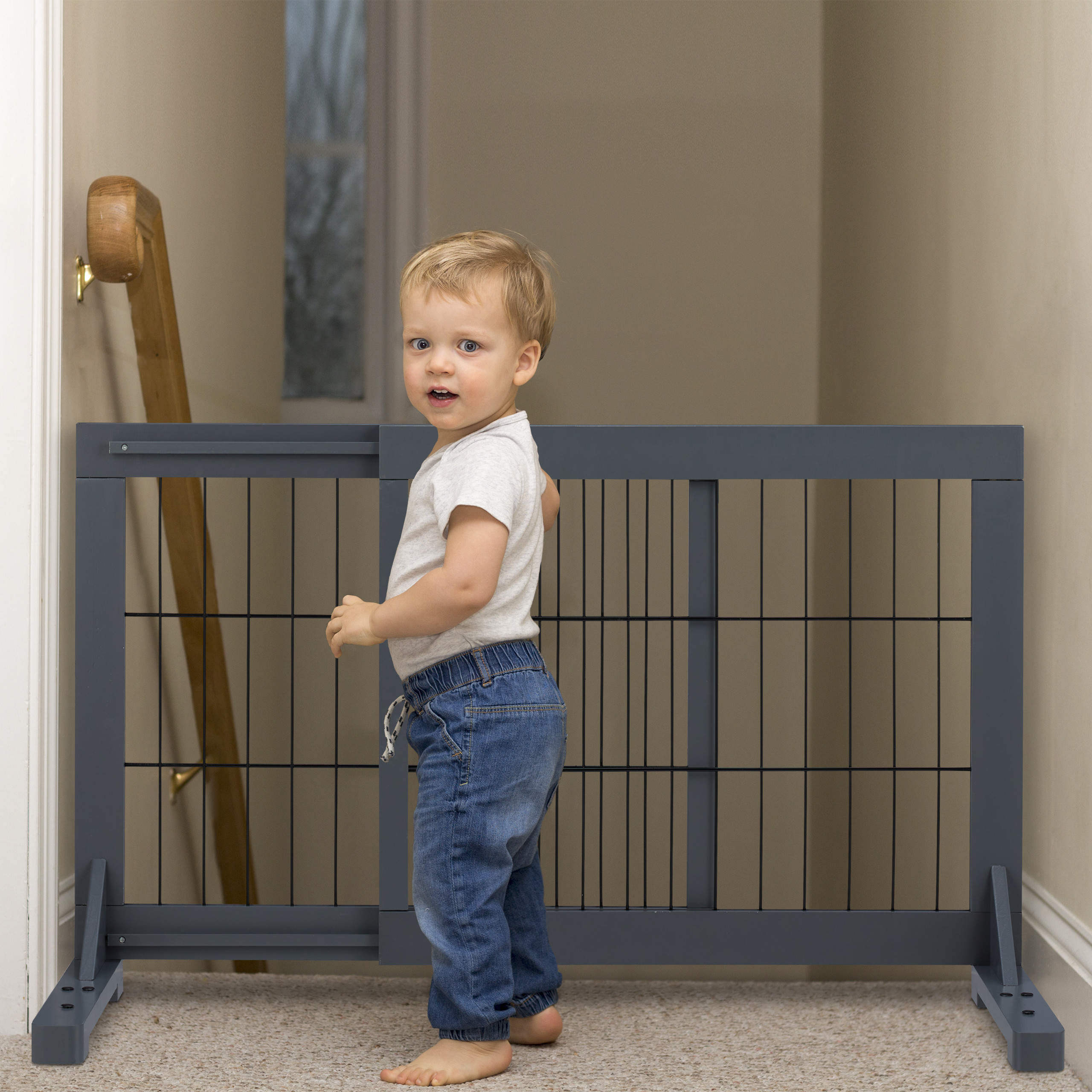Ein Kleinkind in weißem Hemd und blauen Jeans steht vor einem grauen Schutzgitter an einer Treppe.