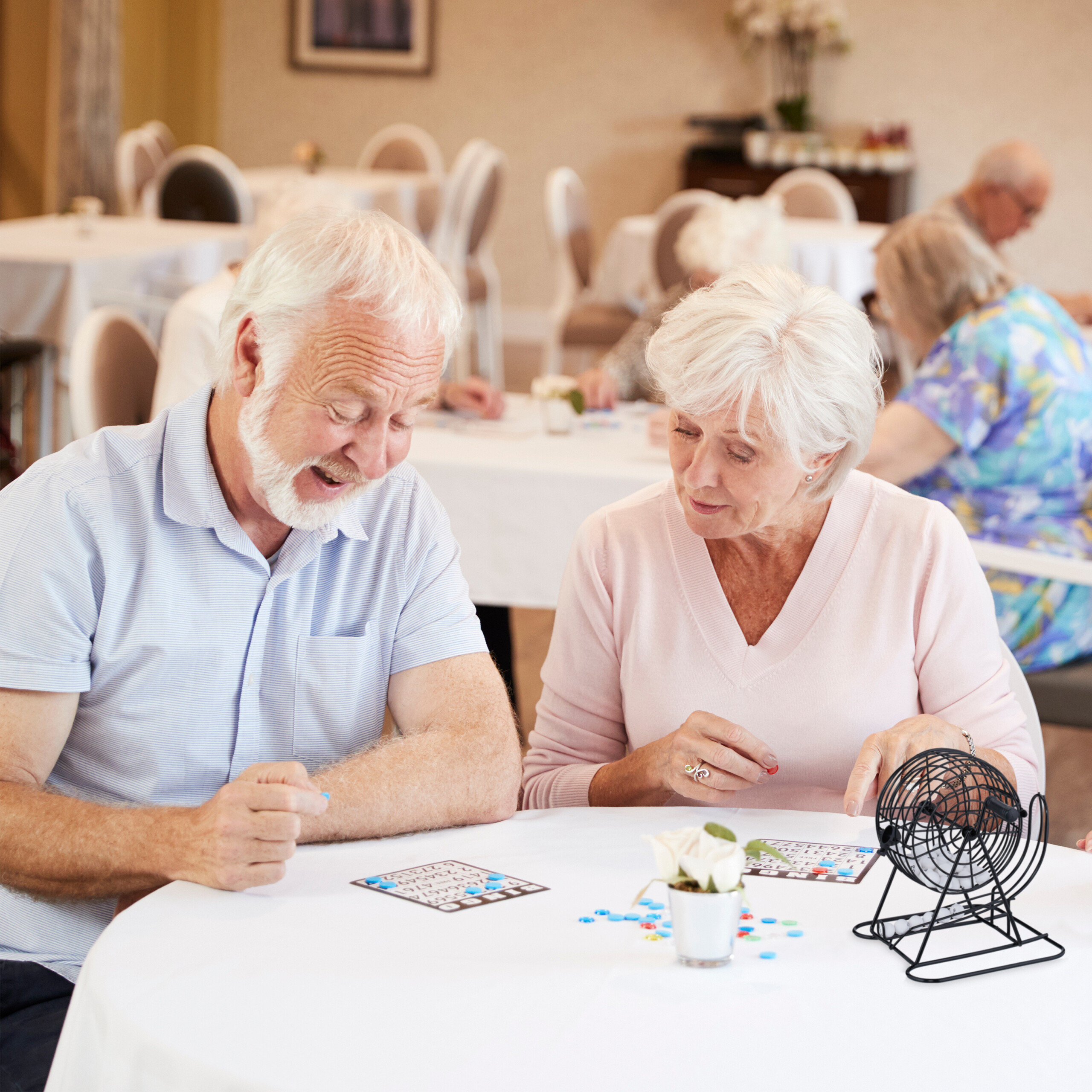 Älteres Paar spielt Bingo an einem weißen Tisch in einem hellen Raum, mit anderen Personen im Hintergrund.