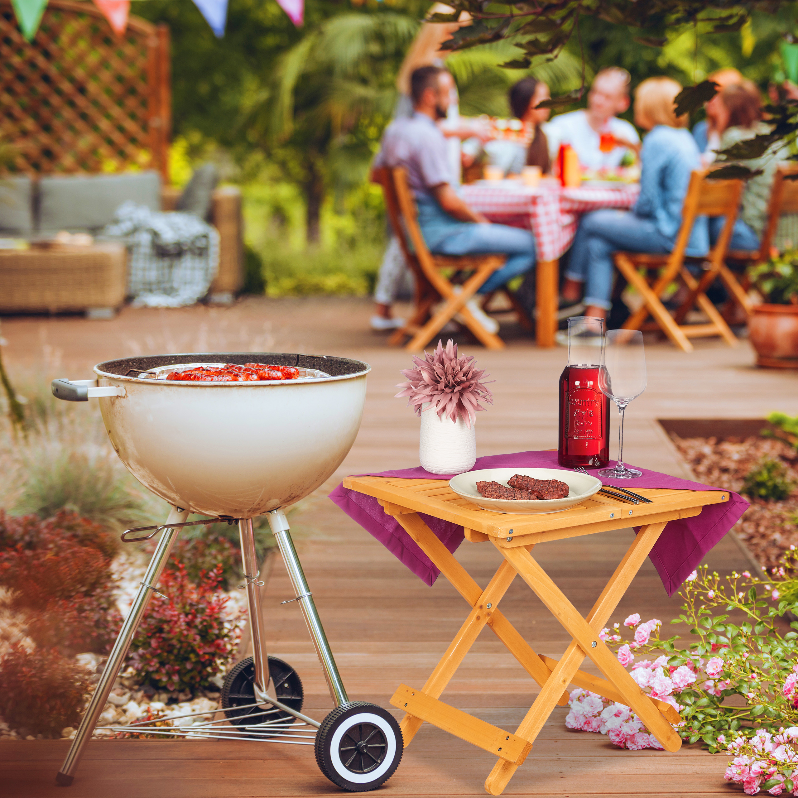 Ein weißer Kugelgrill und ein Holztisch mit rotem Wein und Essen stehen auf einer Holzterrasse; hinten sitzt eine Gruppe.
