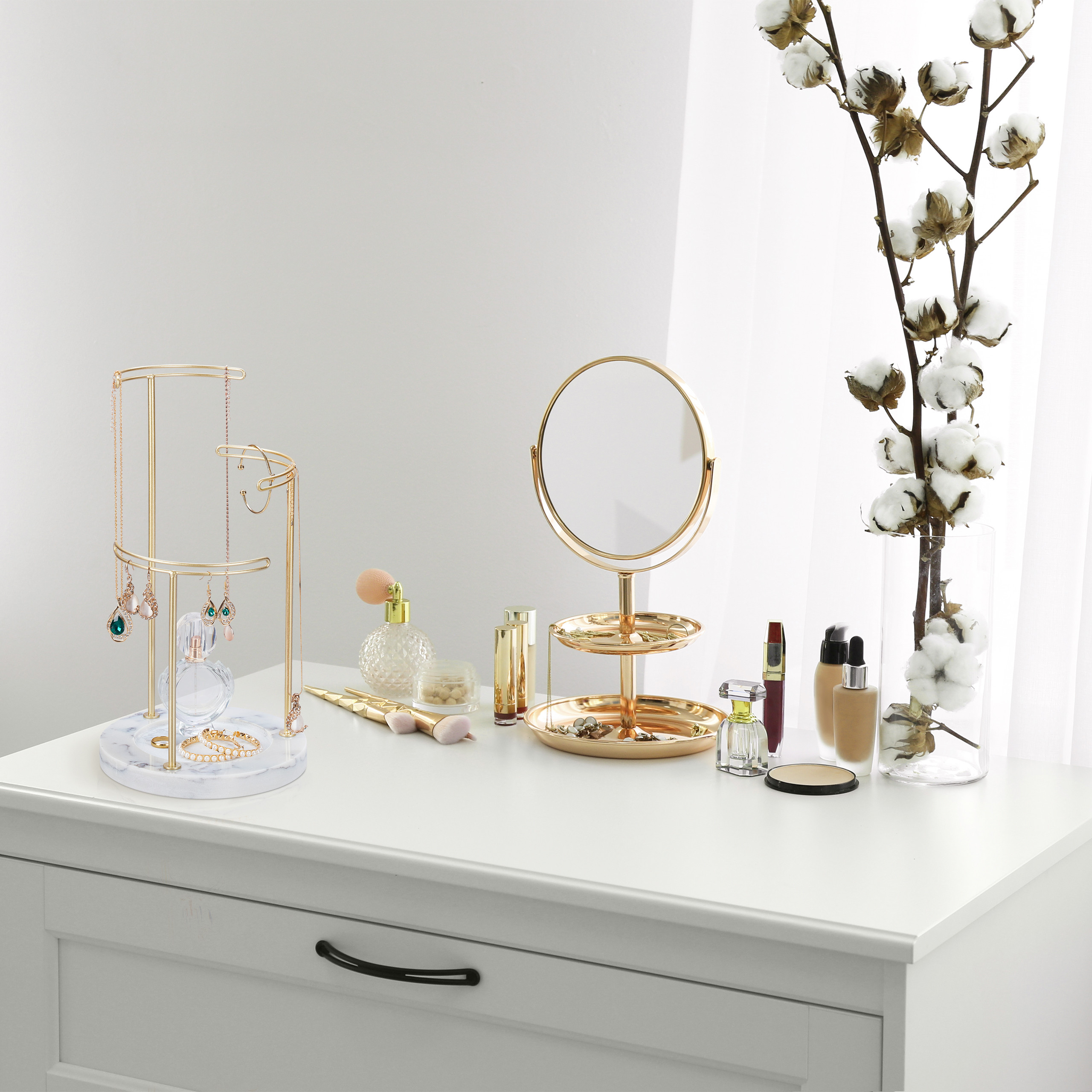 Ein weißer Frisiertisch mit goldenem Schmuckständer, Schminkspiegel, Kosmetik und einer Vase mit Baumwollzweigen.