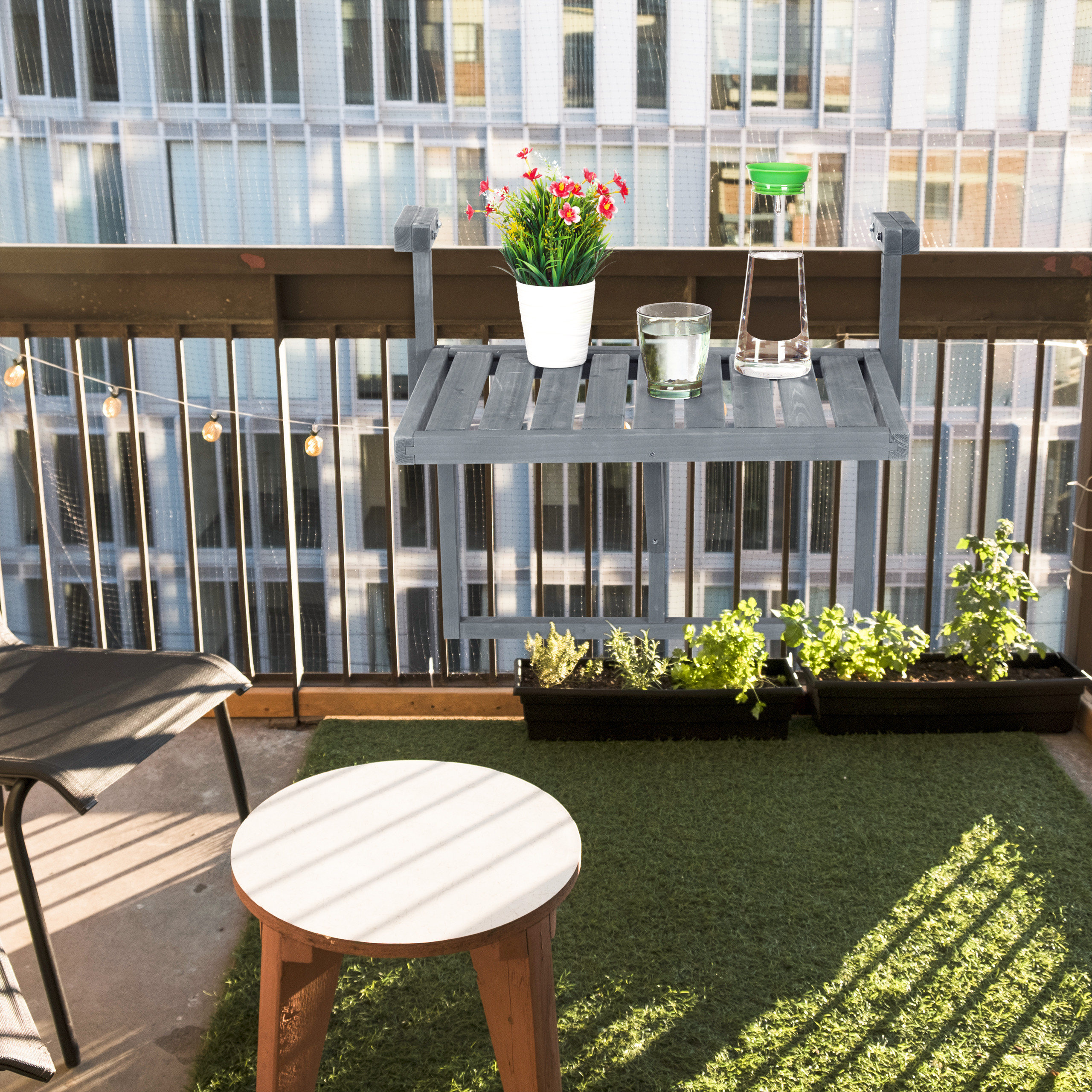 Balkon mit hängendem Tisch, Blumen, Gläsern, einem kleinen Hocker, Kunstrasen und Blick auf das Gebäude.