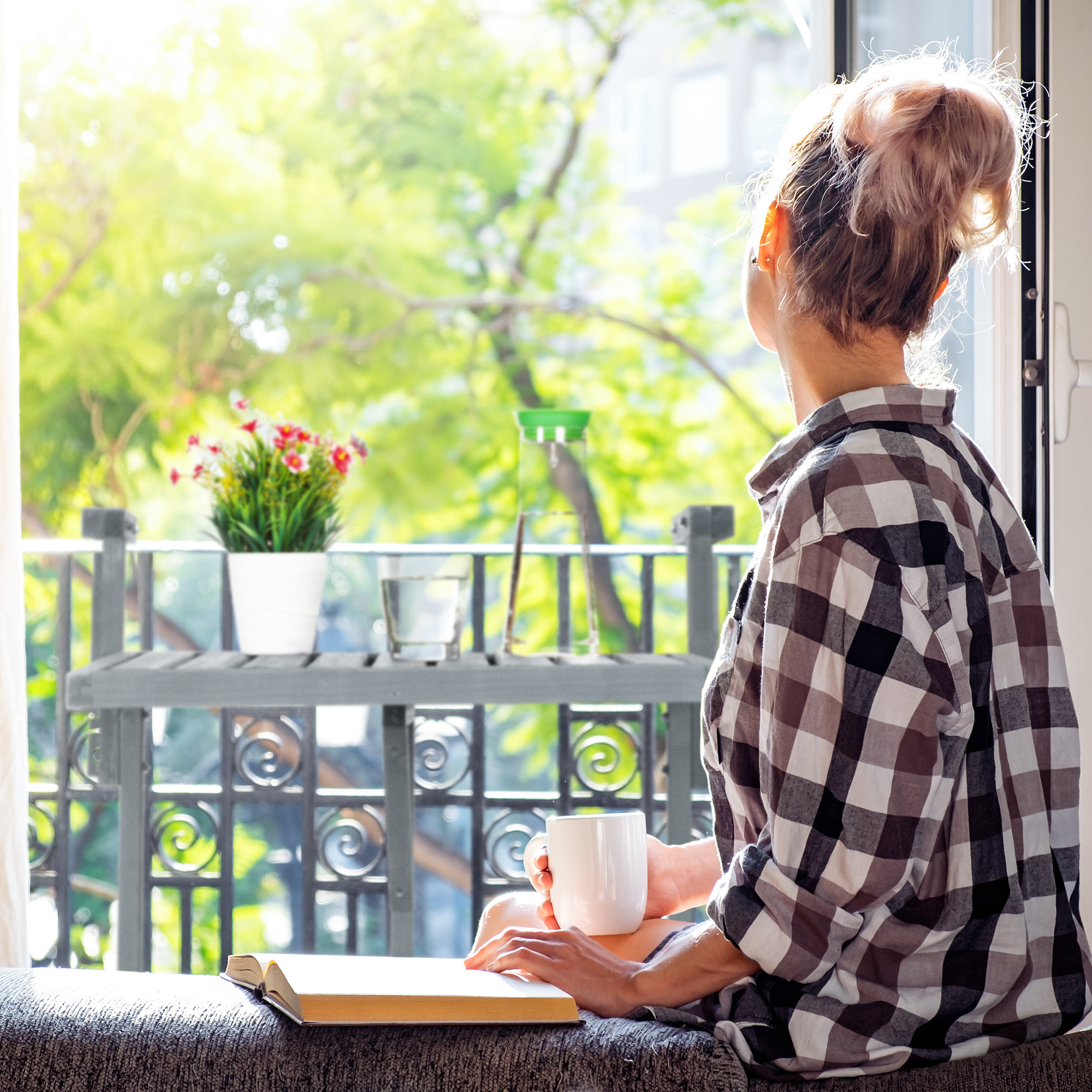 Eine Frau in kariertem Hemd sitzt drinnen mit einer Tasse und einem Buch und blickt auf einen sonnigen Balkon.