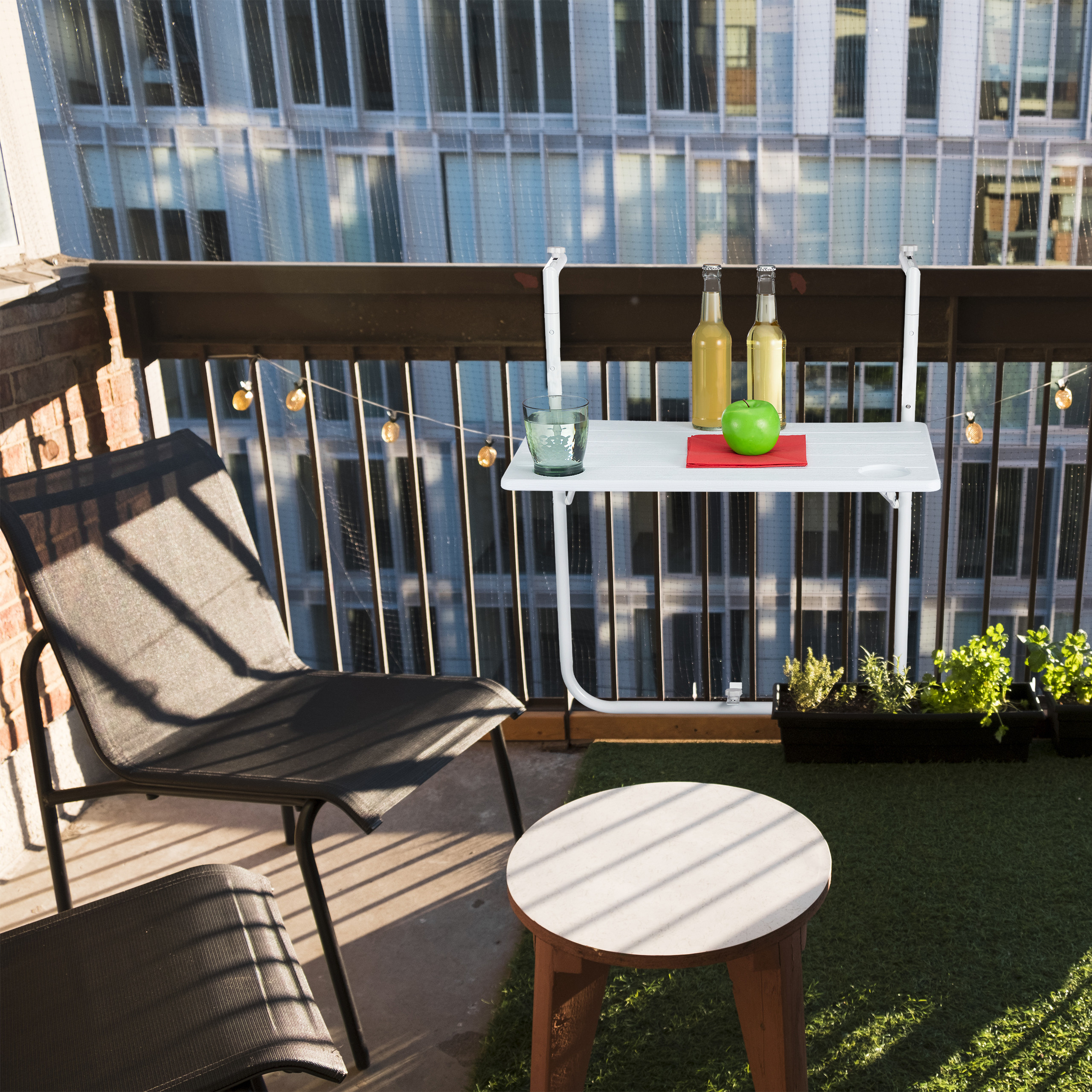 Balkon mit schwarzem Stuhl, weißem Klapptisch mit Flaschen und Apfel sowie künstlichem Rasen vor modernem Gebäude.