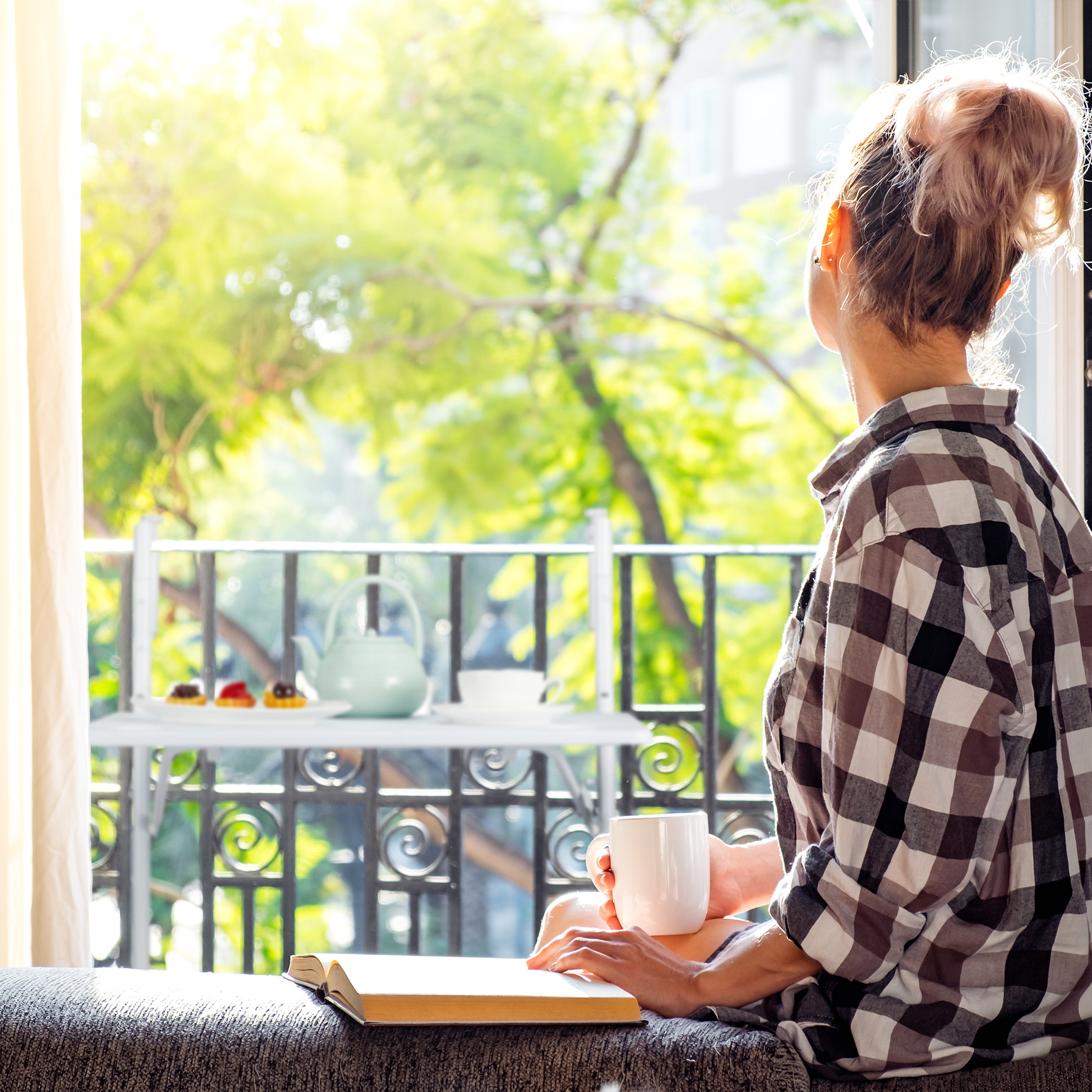 Eine Frau sitzt am offenen Fenster, hält eine Tasse und blickt auf einen sonnigen Balkon.