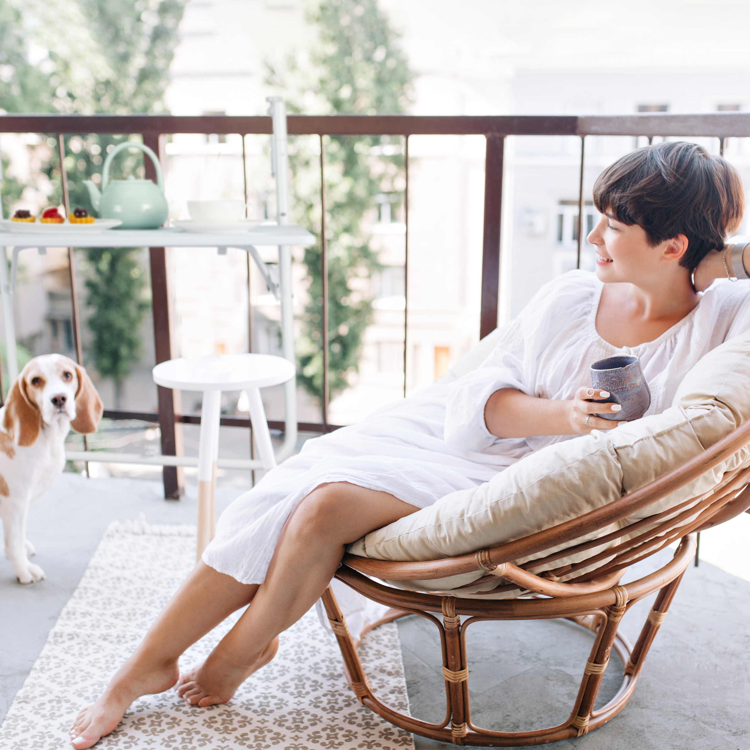 Eine Frau im weißen Kleid sitzt auf einem Balkon in einem Rattan-Sessel mit Tasse, während ein Beagle zusieht.