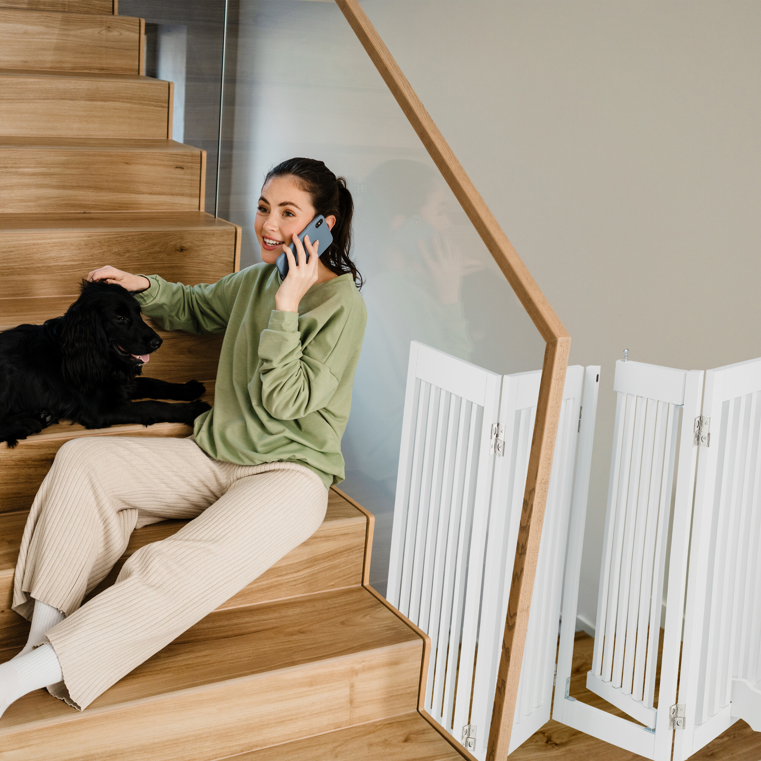 Eine Frau in einem grünen Pullover sitzt auf einer Holztreppe, streichelt einen schwarzen Hund und telefoniert.