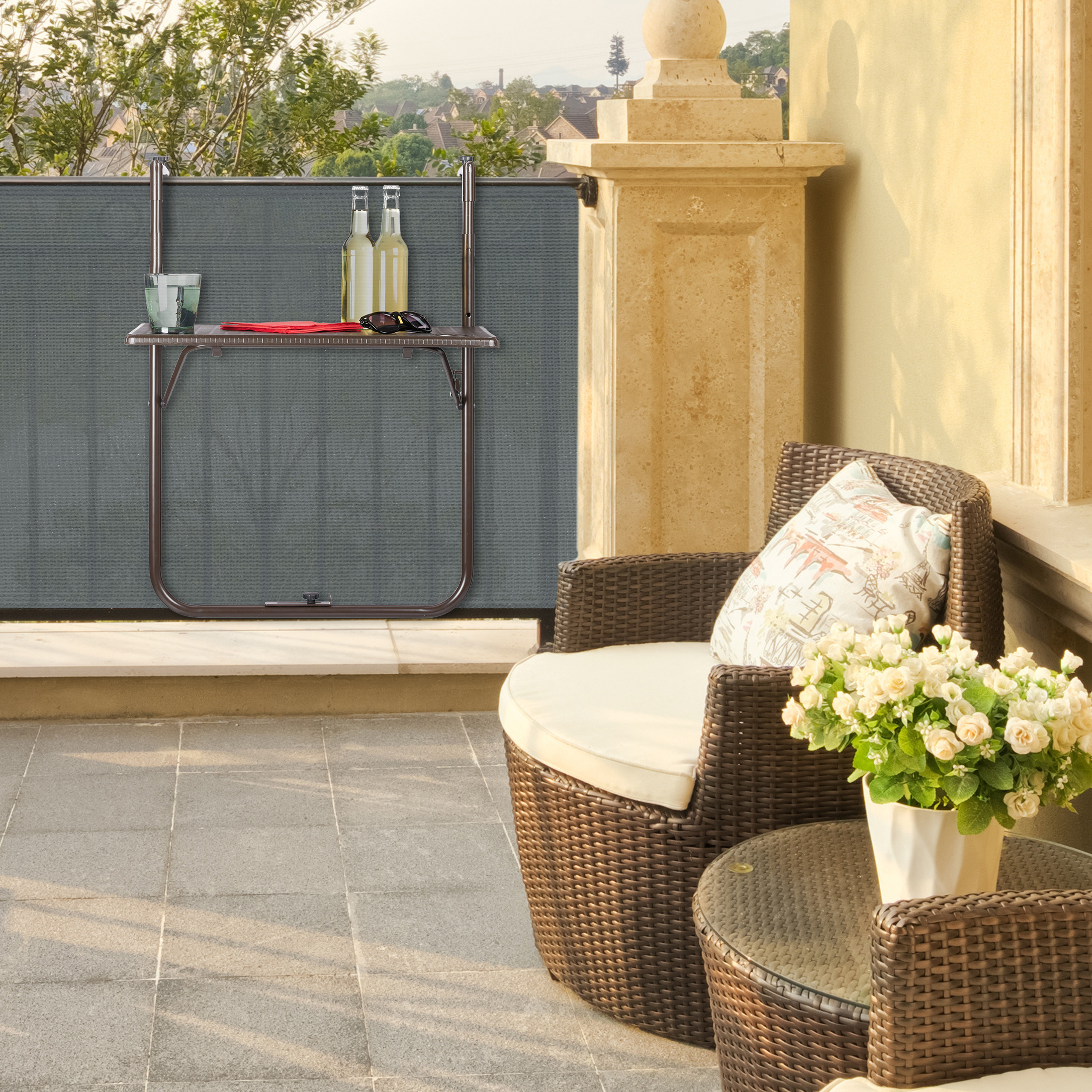 Ein Balkon mit braunem Korbstuhl, kleiner Tisch mit weißen Blumen und klappbarem Tisch am Geländer.