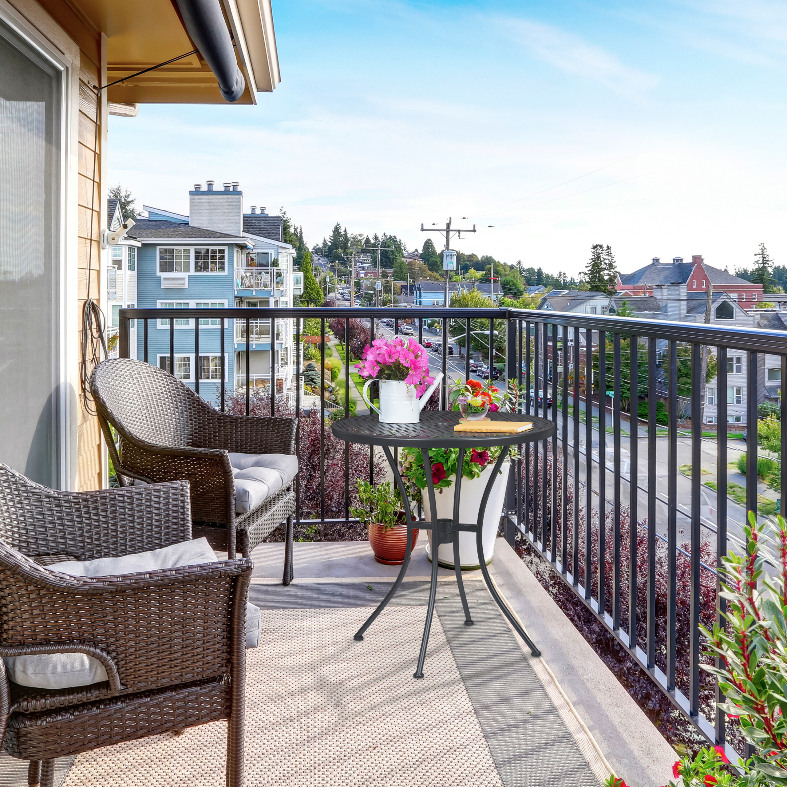 Ein sonniger Balkon mit zwei Rattansesseln, einem Tisch mit Blumen und Blick auf Häuser und Bäume.