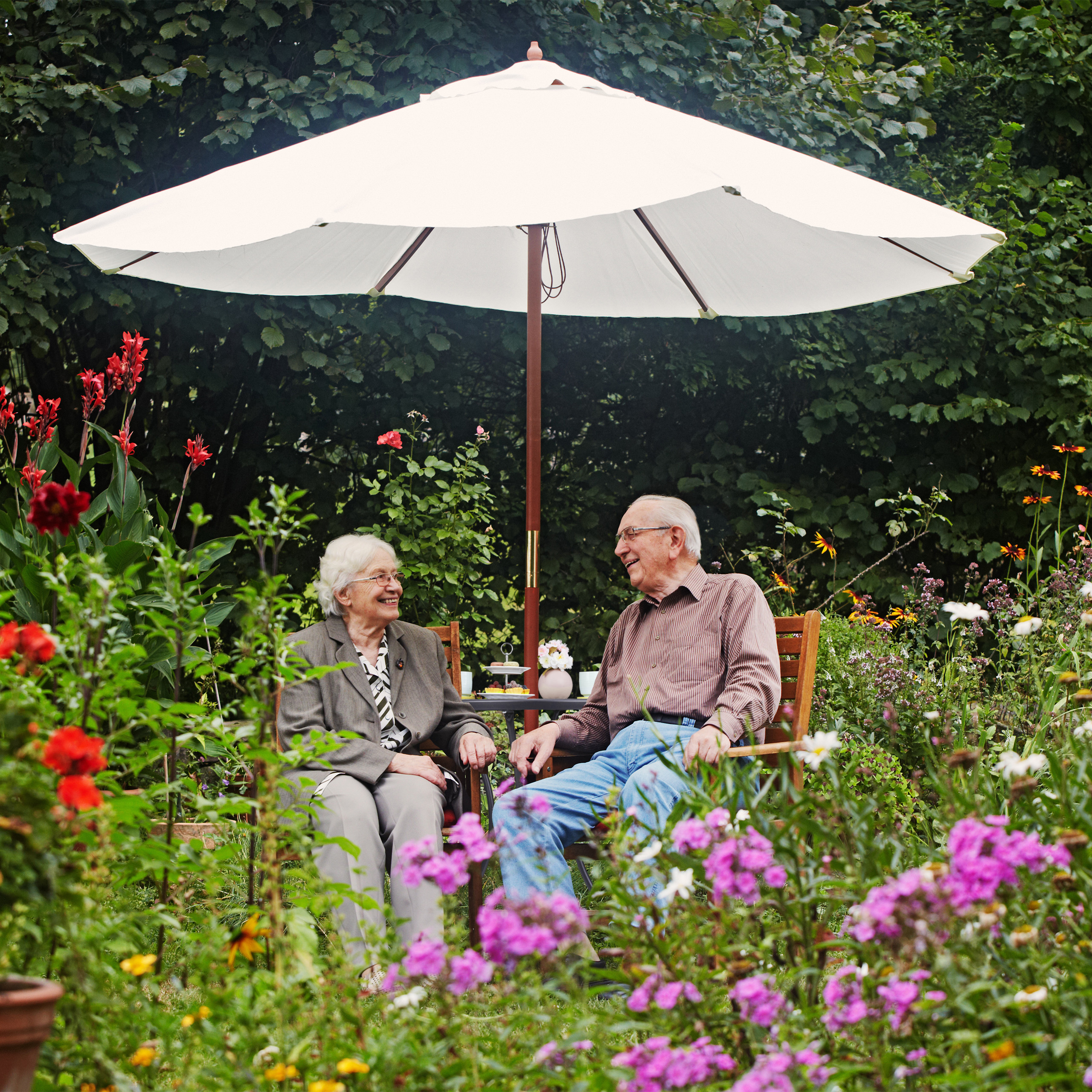 Älteres Paar sitzt lachend unter einem weißen Sonnenschirm in einem blühenden Garten.