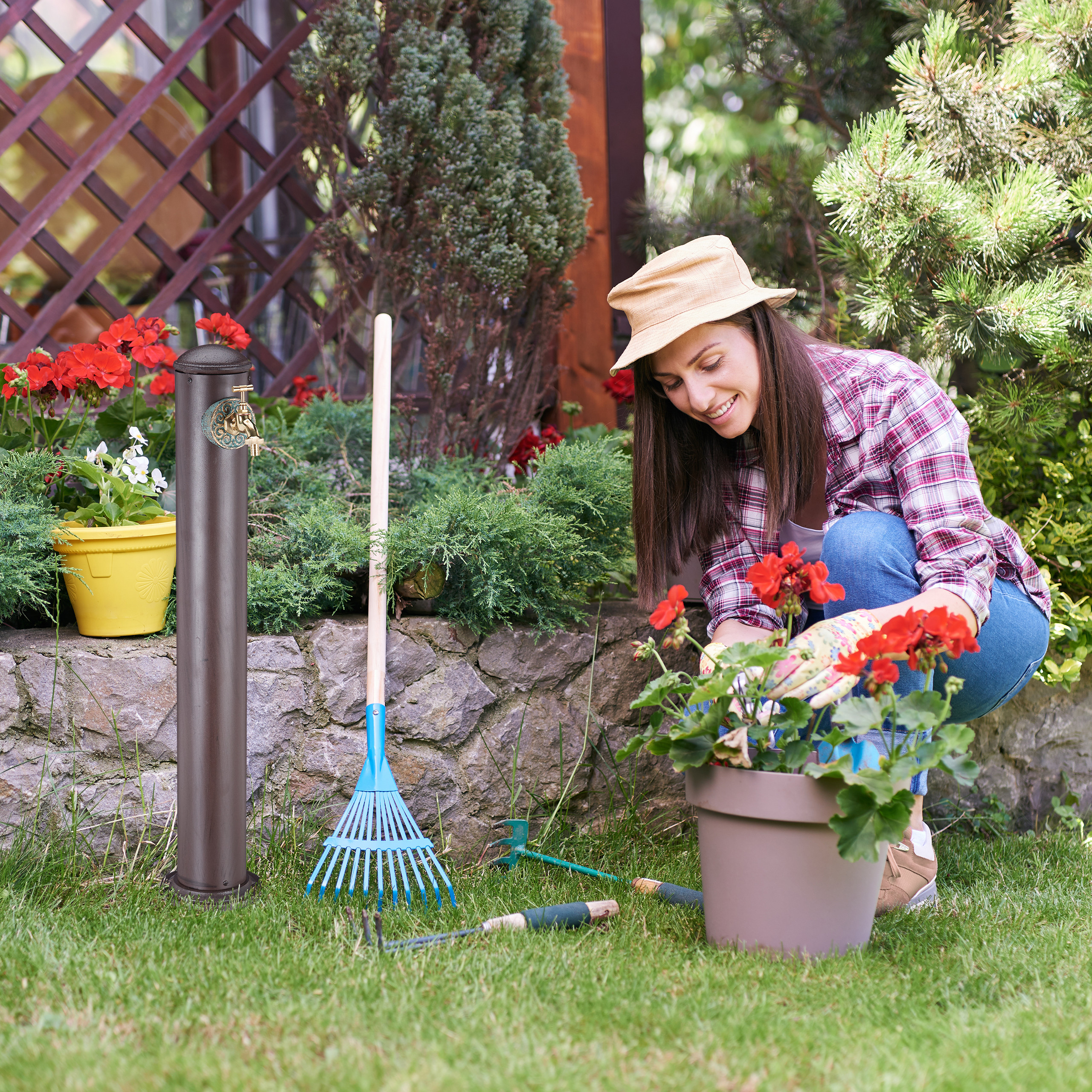 Eine Frau mit Hut pflegt rote Blumen in einem Topf im Garten, neben einer Säule und Gartengeräten.