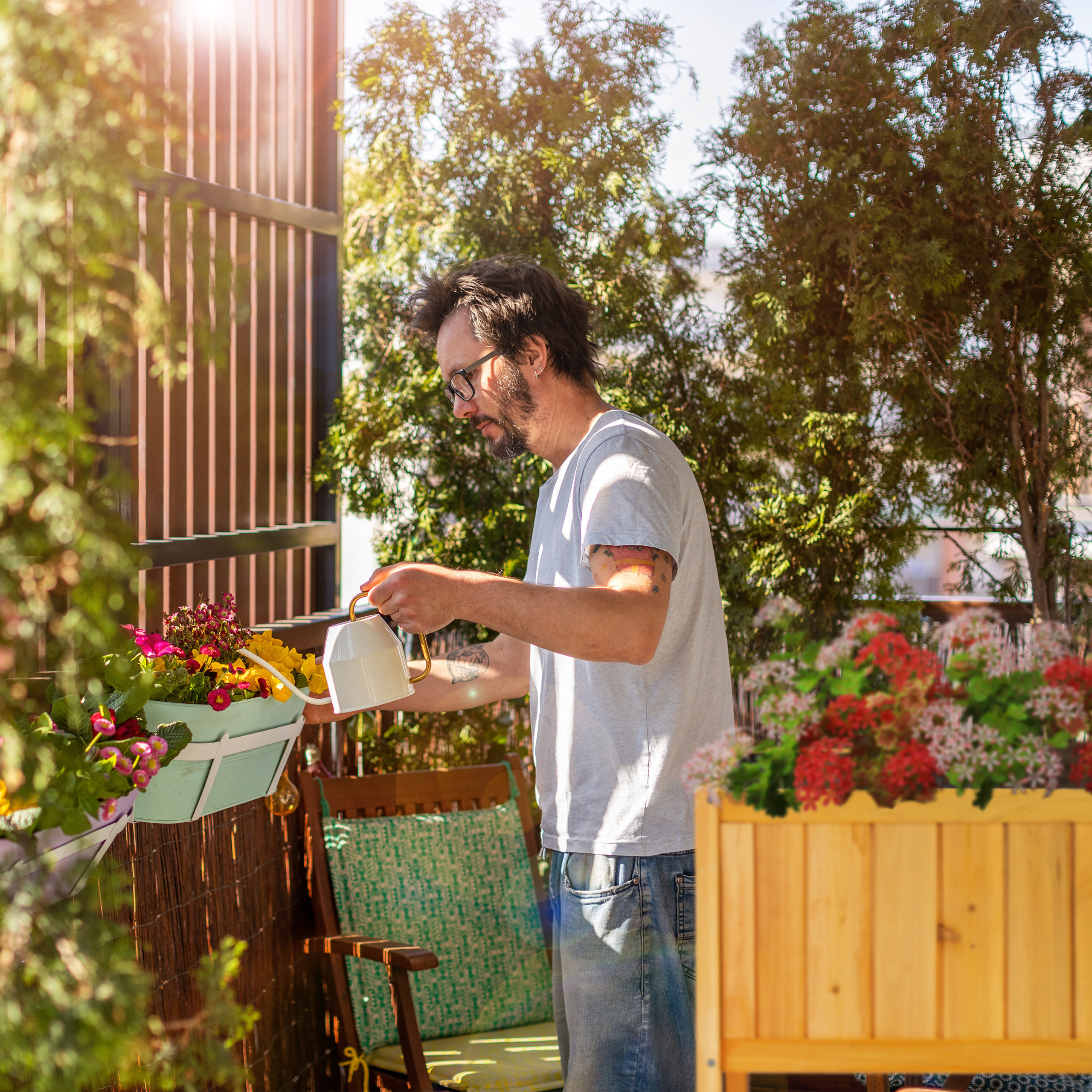 Ein Mann mit Brille gießt bunte Blumen in Töpfen auf einem sonnigen Balkon mit üppigen grünen Bäumen.