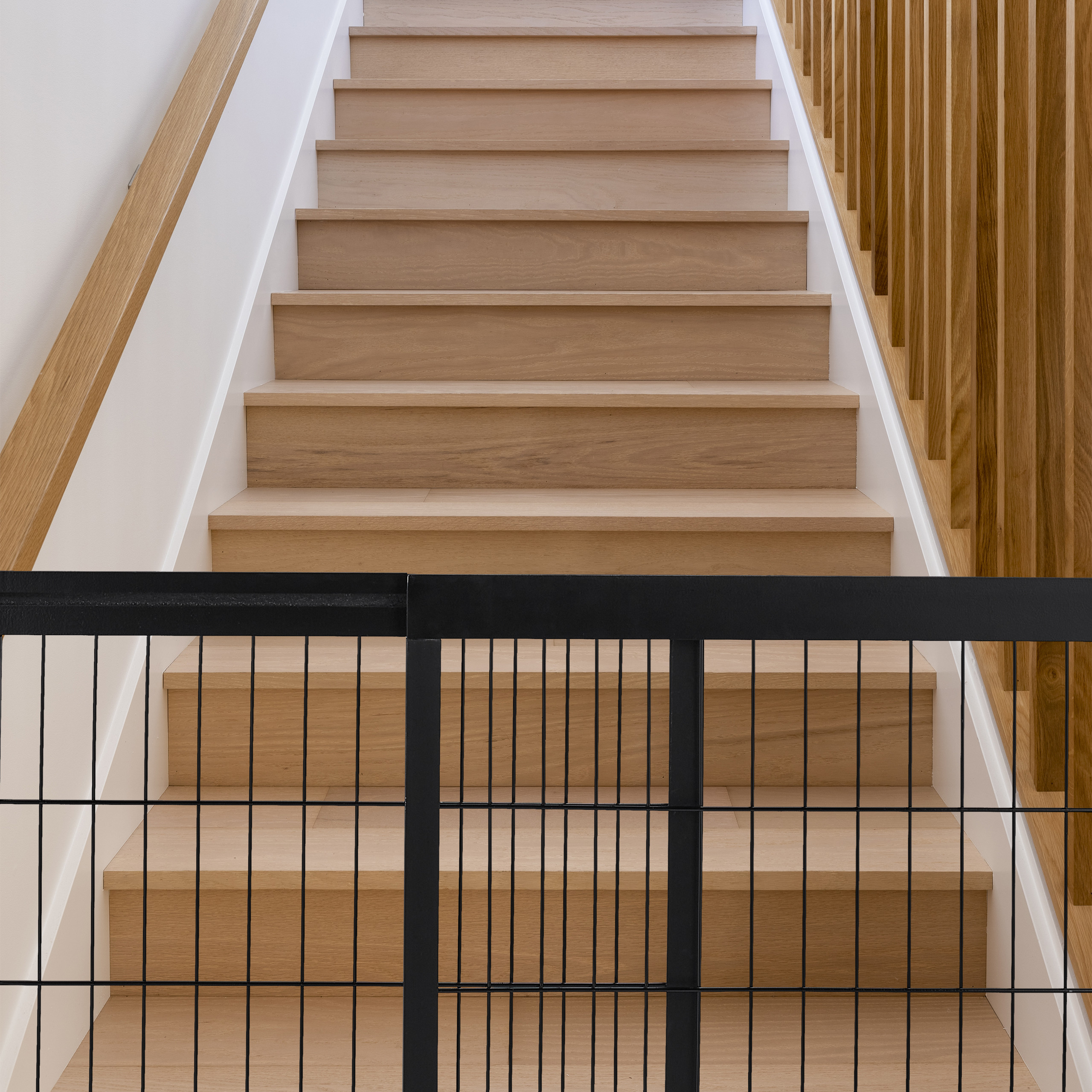 Holztreppe mit weißer Wand, Holzbalkengeländer und einem schwarzen Schutzgitter unten.