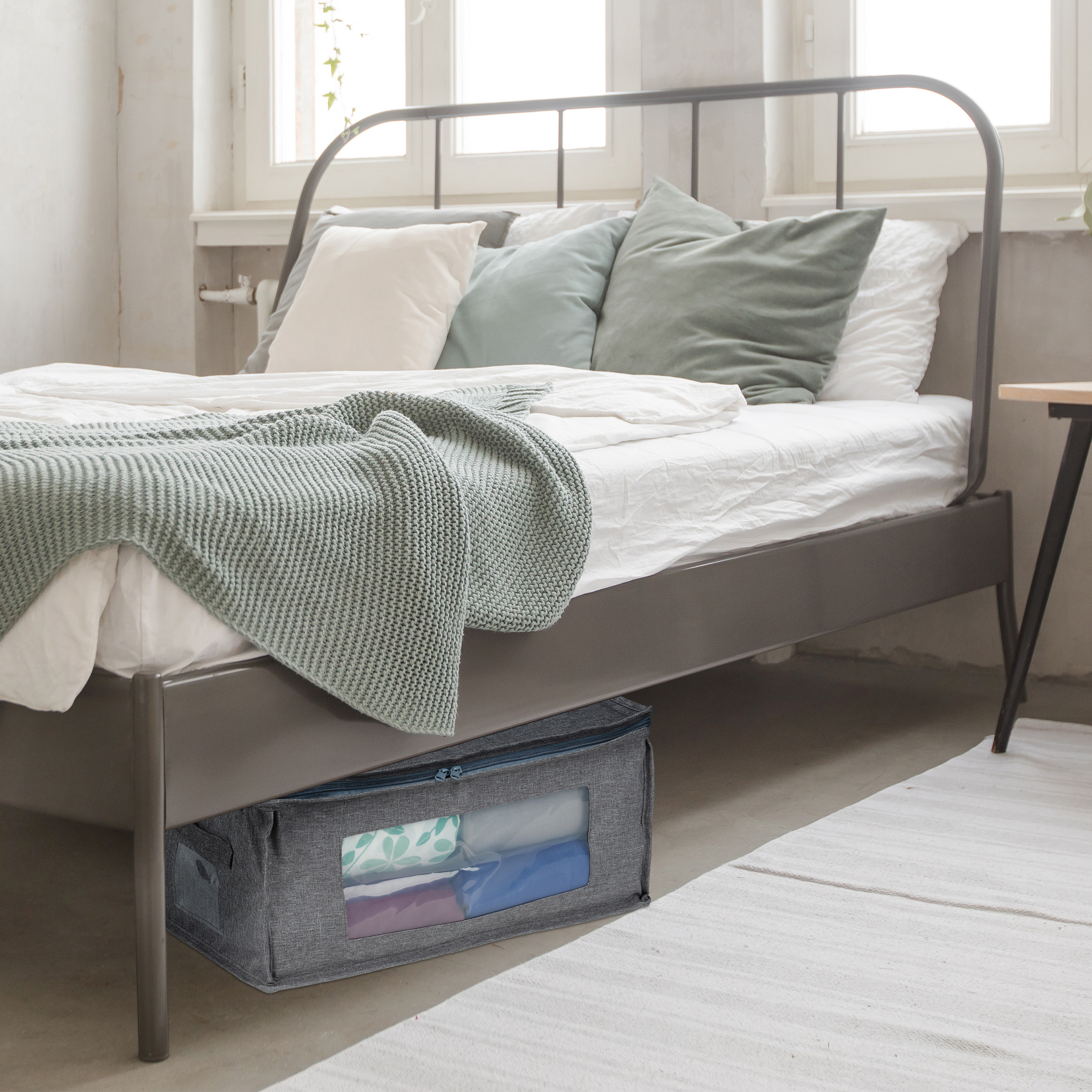 Ein grauer Stoff-Aufbewahrungsbehälter mit klarem Sichtfenster steht unter einem metallenen Doppelbett in einem hellen Zimmer.