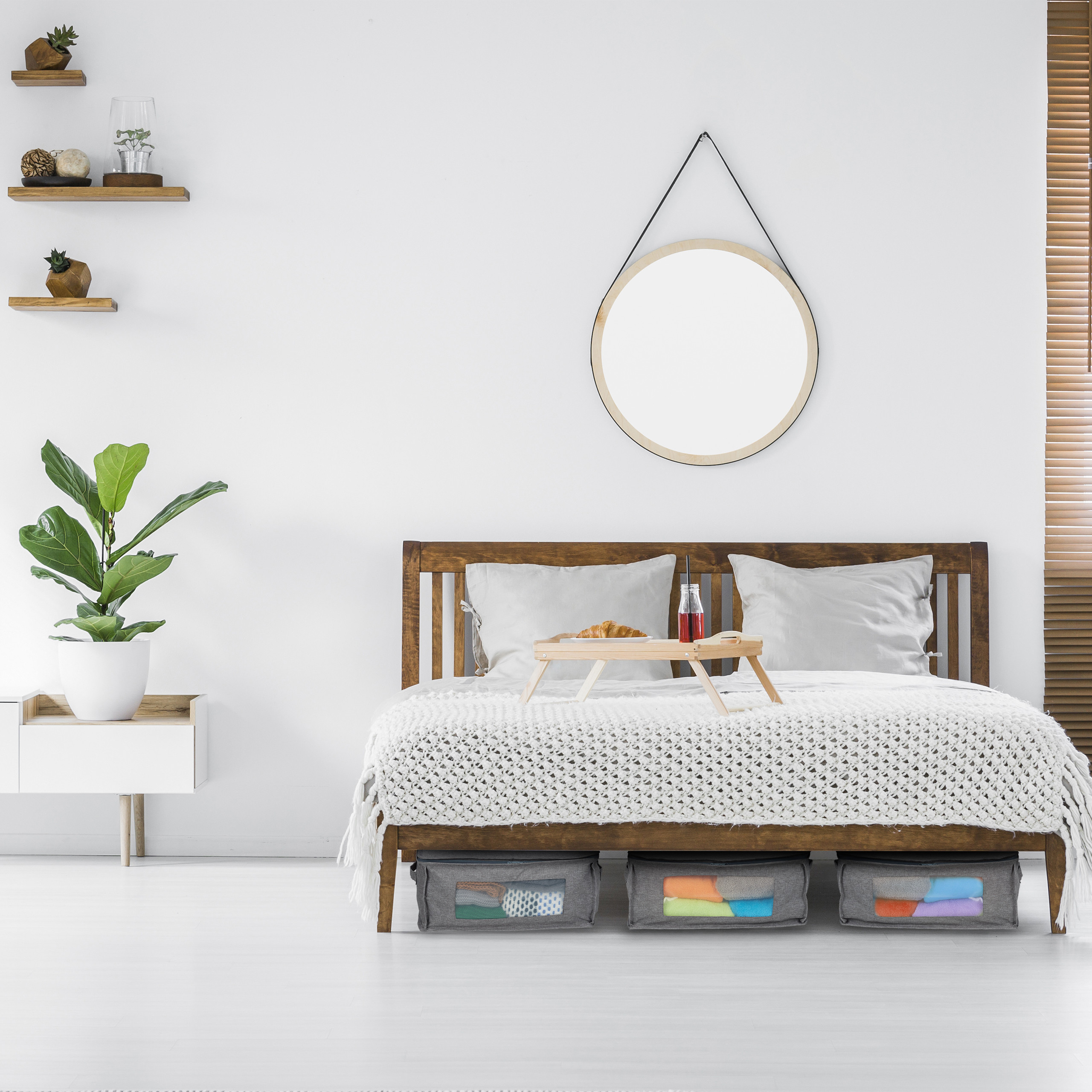 Ein Holzbett mit grauen Kissen, weißer Decke, Tablett, Aufbewahrungsboxen darunter, rundem Spiegel und Regalen.