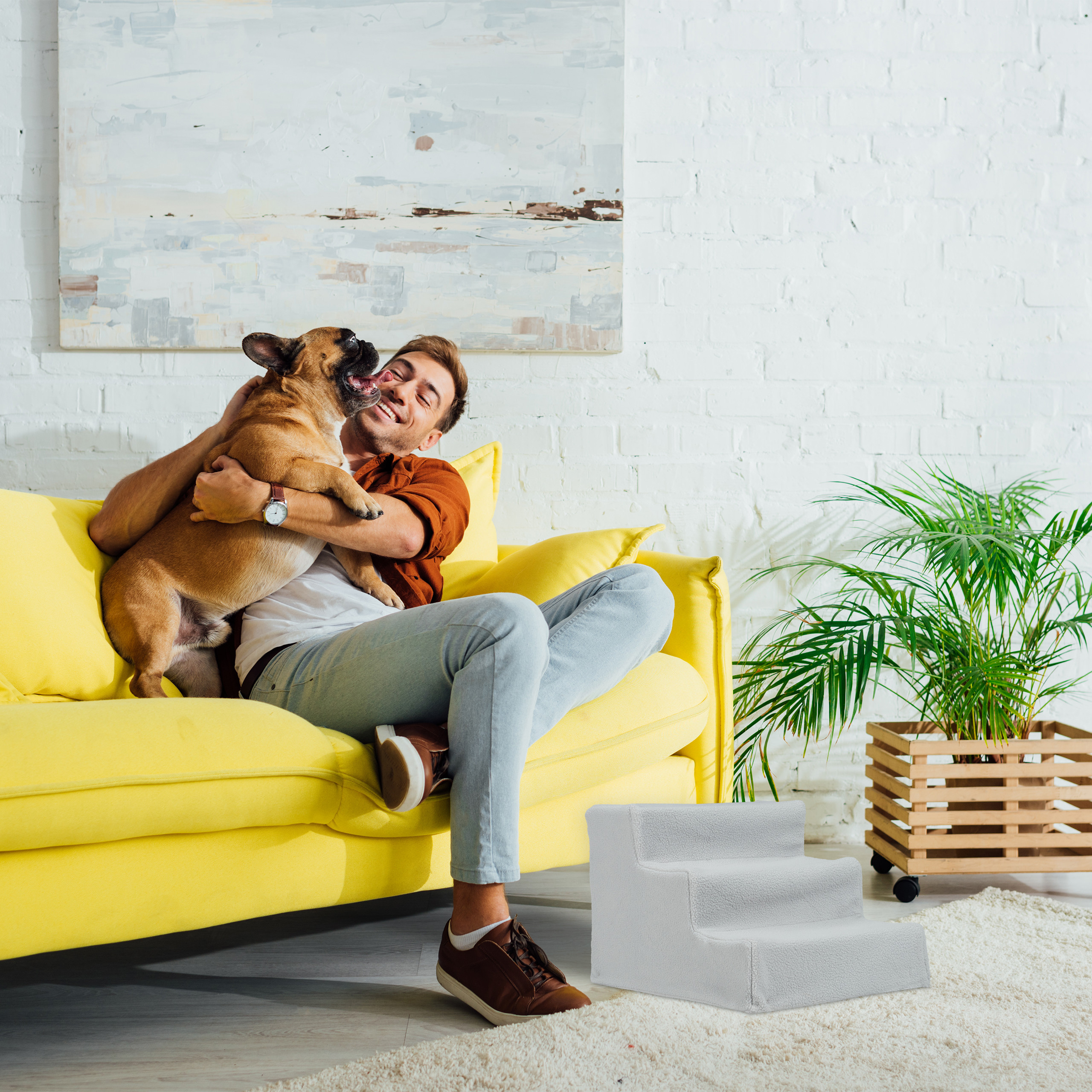 Ein glücklicher Mann kuschelt auf einem gelben Sofa mit seiner französischen Bulldogge in einem hellen Raum.