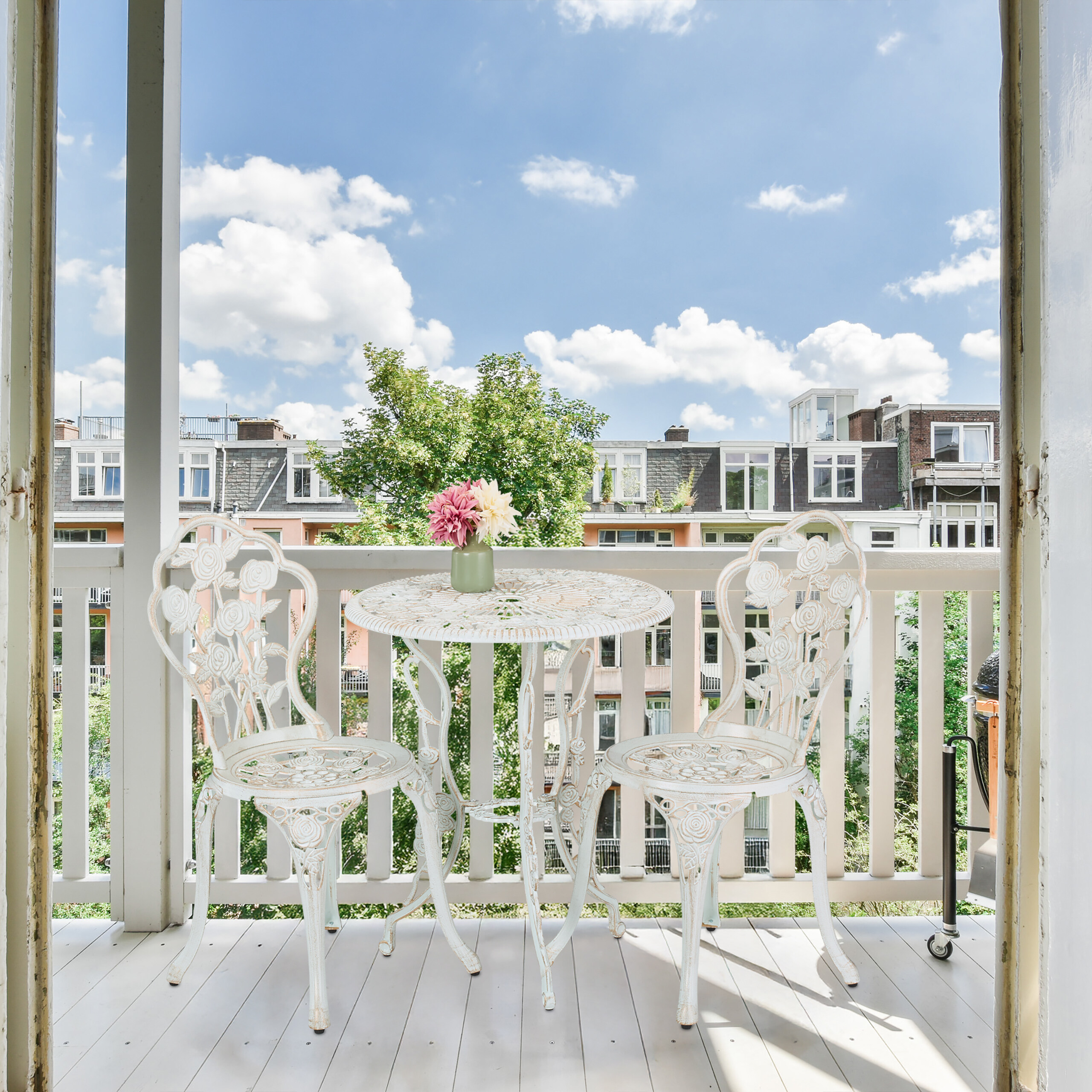 Ein weißer Bistrotisch aus Metall und zwei Stühle mit Blumenmuster auf einem sonnigen Balkon.