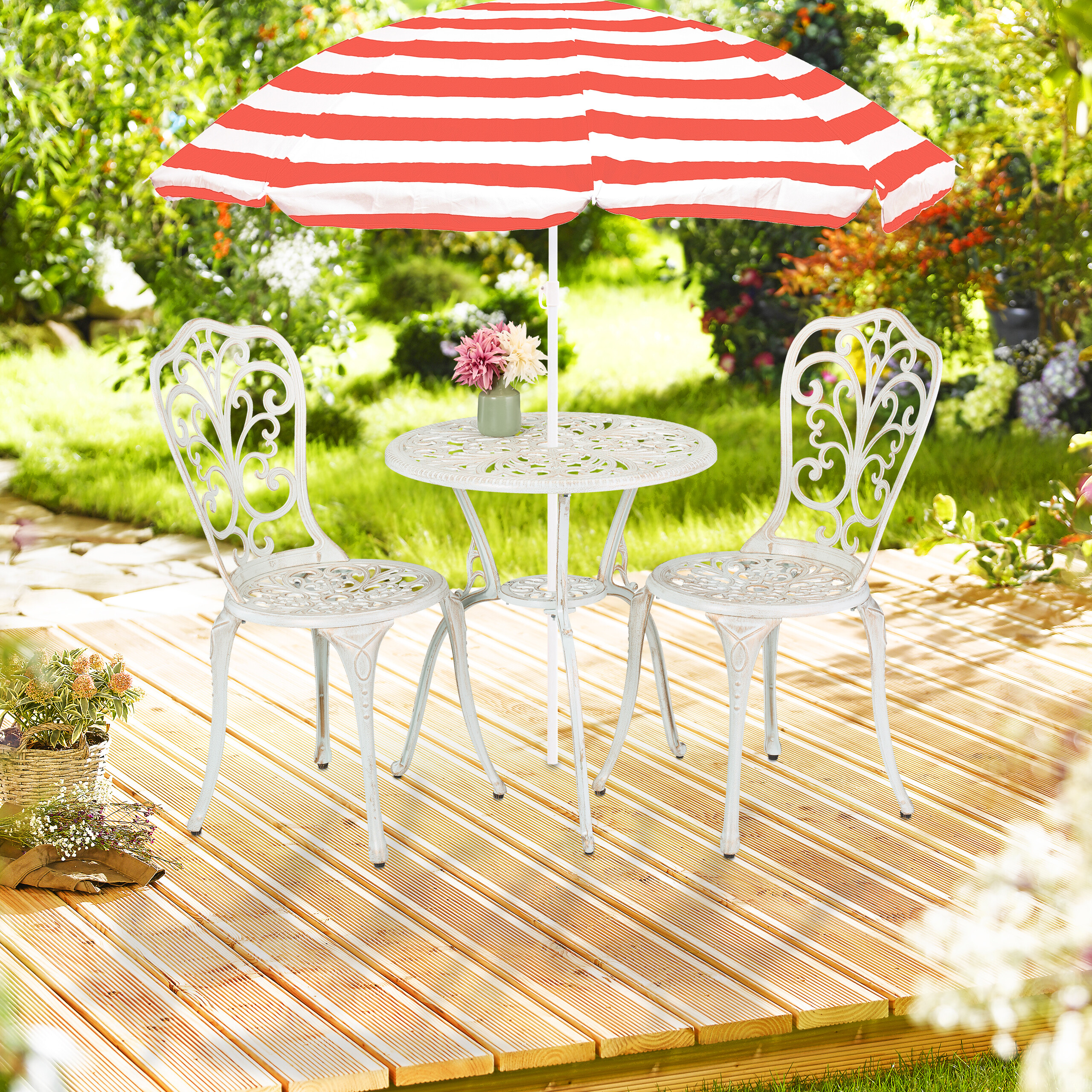 Ein weißer Metallgartentisch und zwei Stühle mit rot-weiß gestreifter Sonnenschirm auf Holzdielen im Garten.