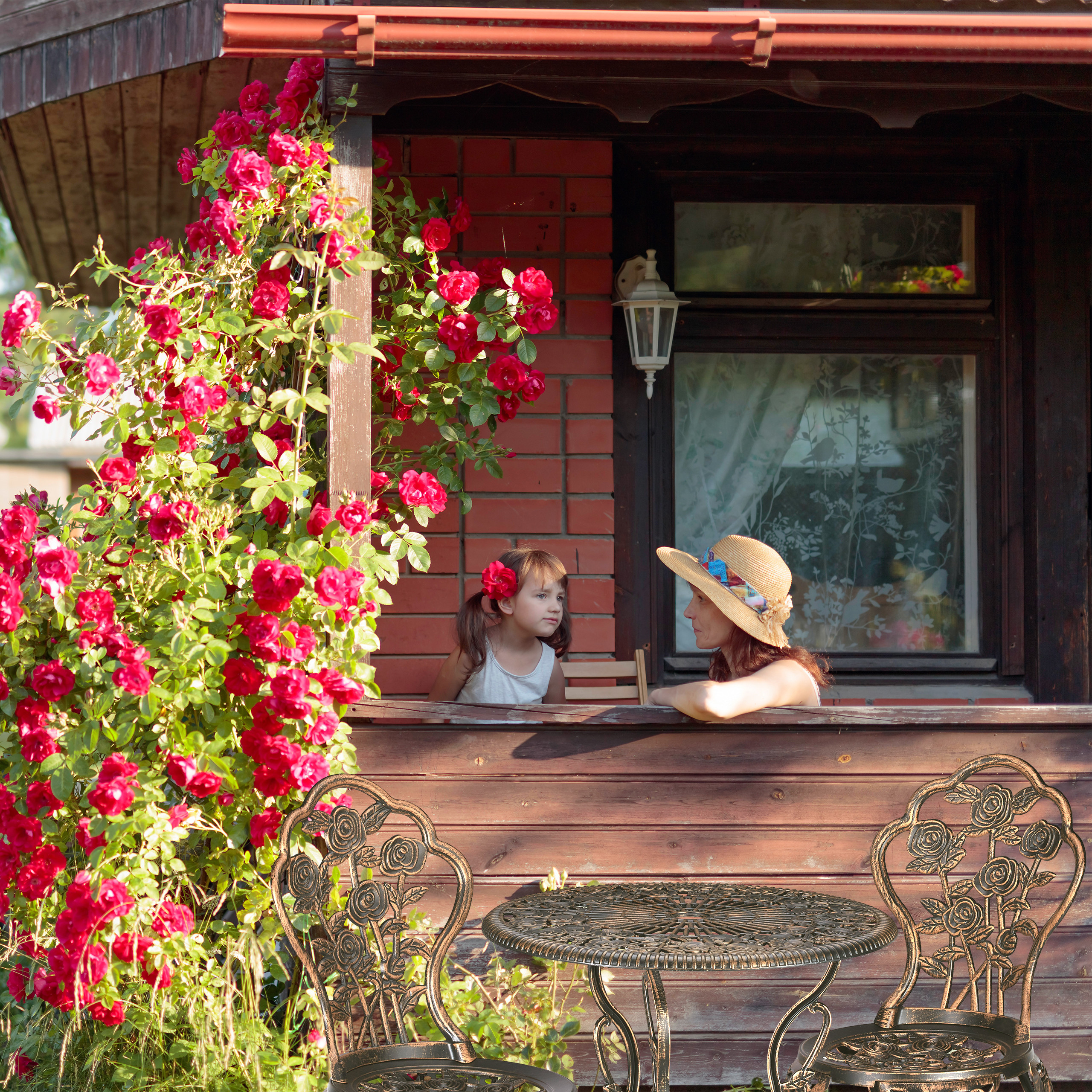 Eine Frau und ein Mädchen sitzen auf einer Holzveranda neben einem üppigen Busch voller roter Rosen.