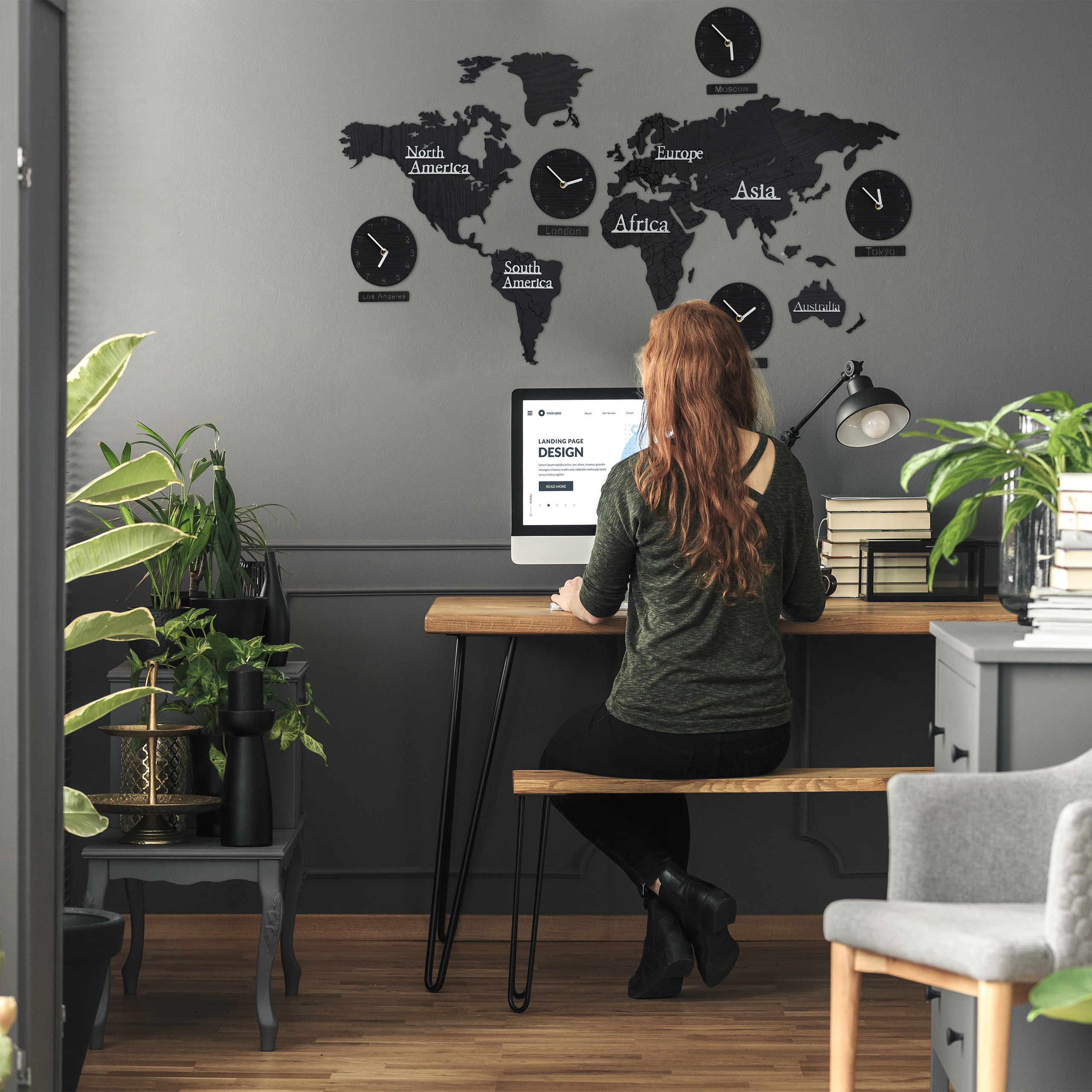 Eine Frau arbeitet an einem Schreibtisch vor einer grauen Wand mit Weltkarte und Uhren.