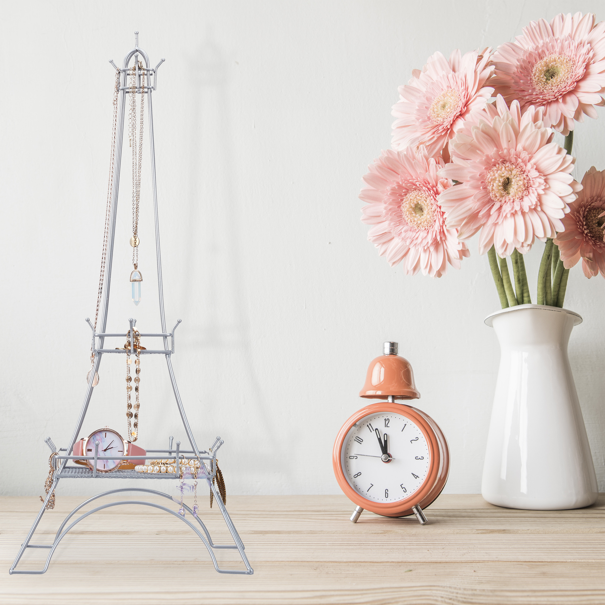Ein Eiffelturm-Schmuckhalter, eine orangefarbene Weckuhr und eine weiße Vase mit rosa Blumen auf Holz.