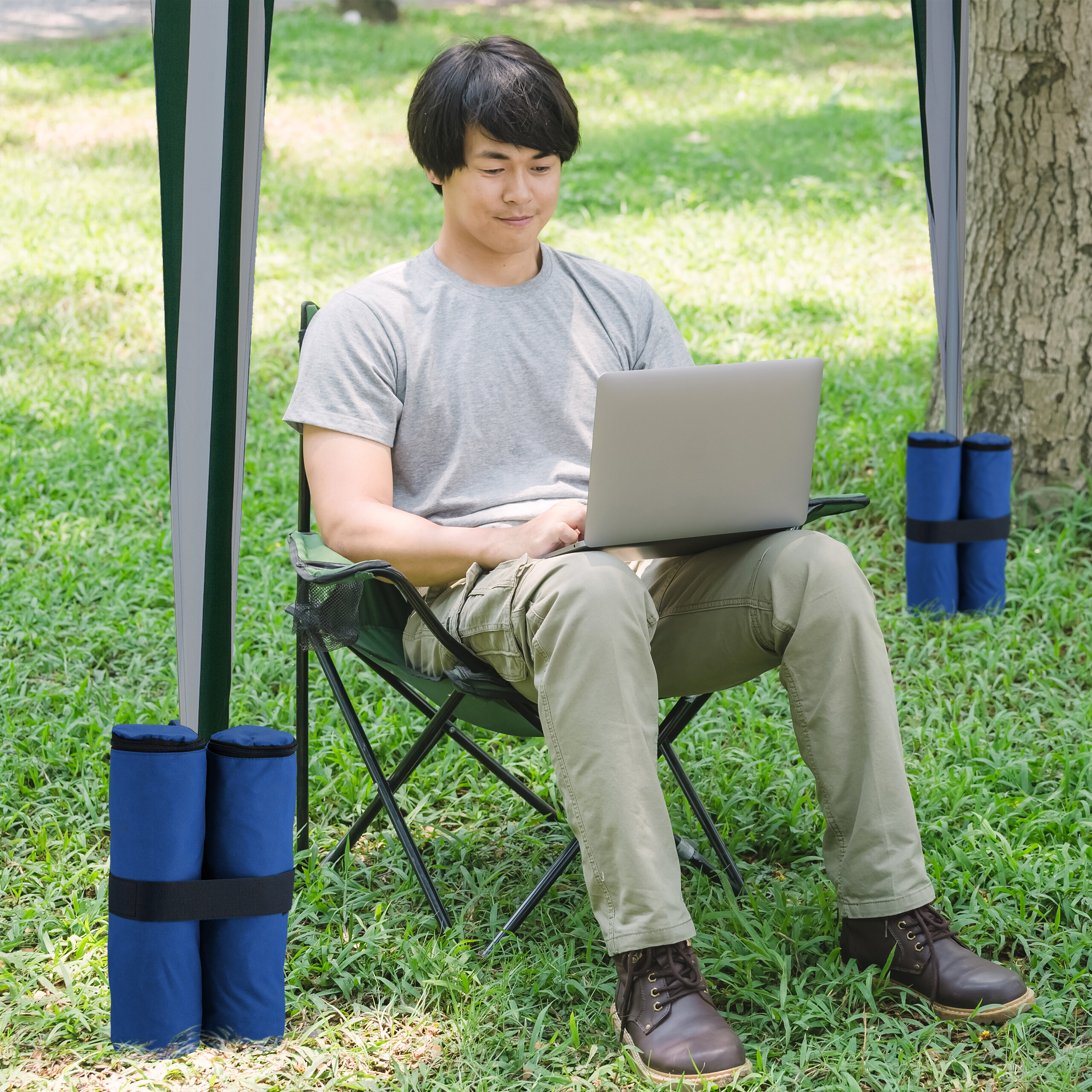 Ein Mann sitzt in einem Campingstuhl im Gras und arbeitet an einem Laptop unter einem Pavillon.