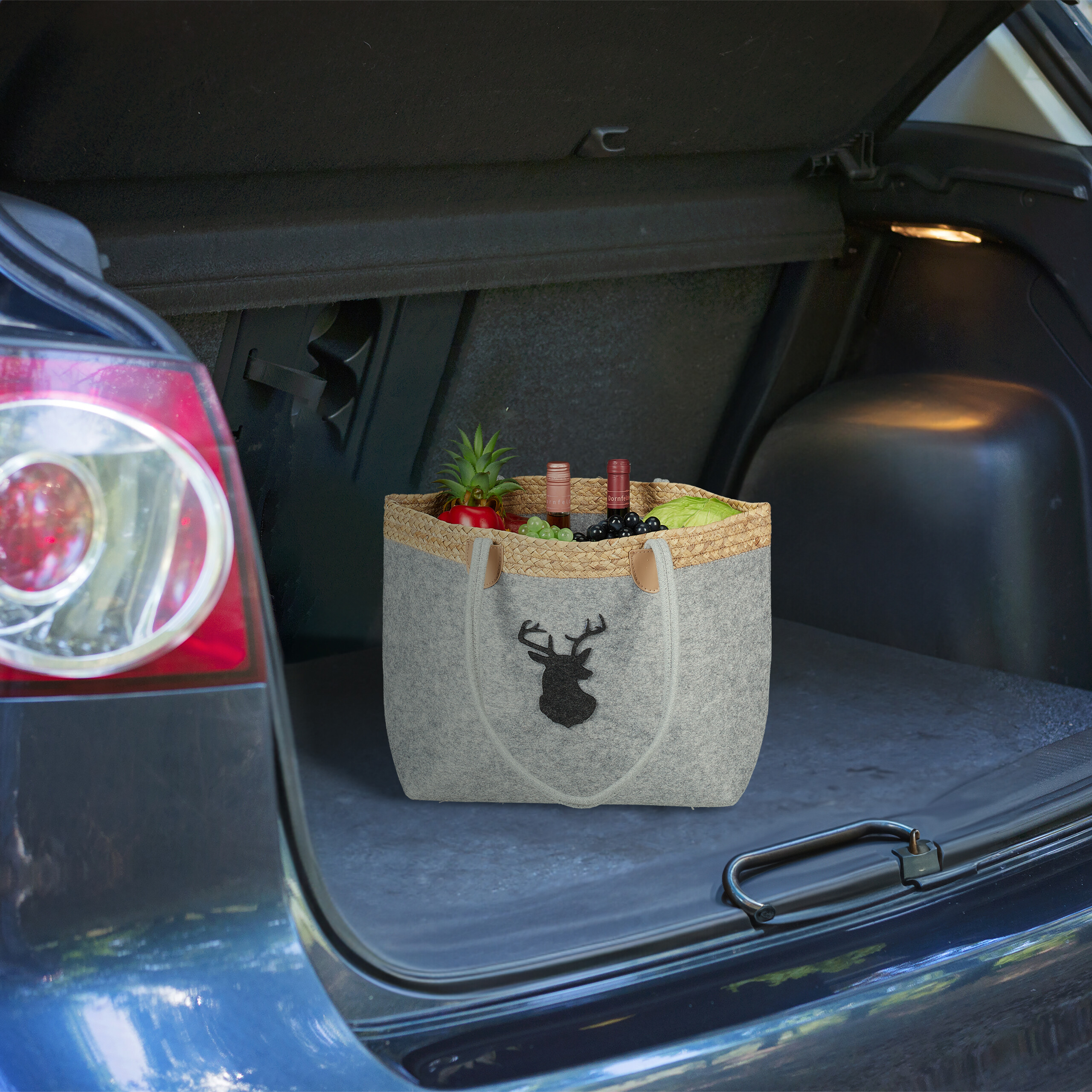 Eine graue Filztasche mit Hirschmotiv und Lebensmitteln steht im offenen Kofferraum eines Autos.
