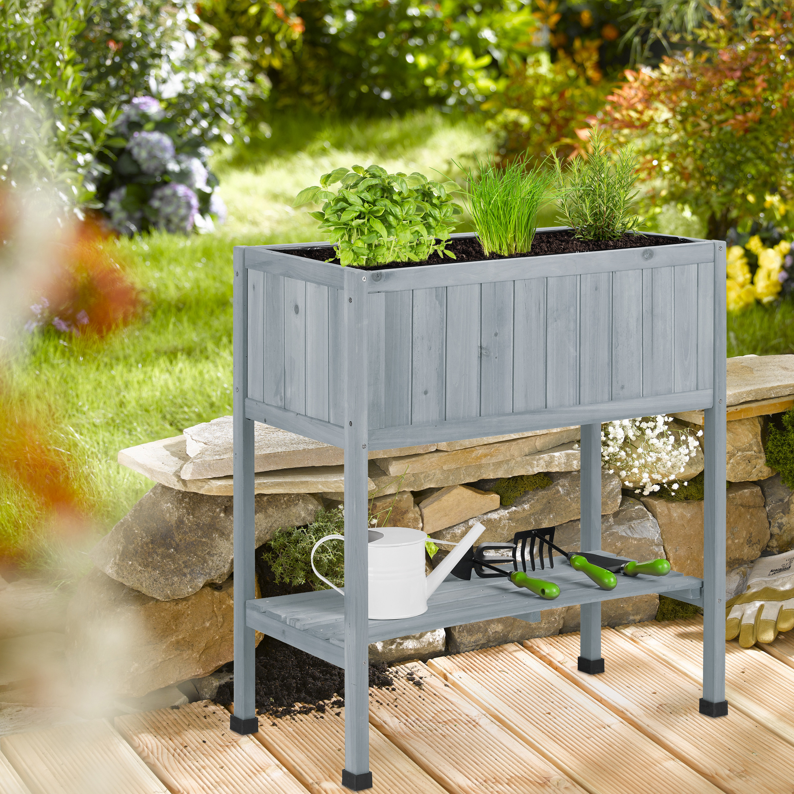 Graues Hochbeet aus Holz mit Kräutern auf Terrasse, darunter eine Gießkanne und Werkzeuge.