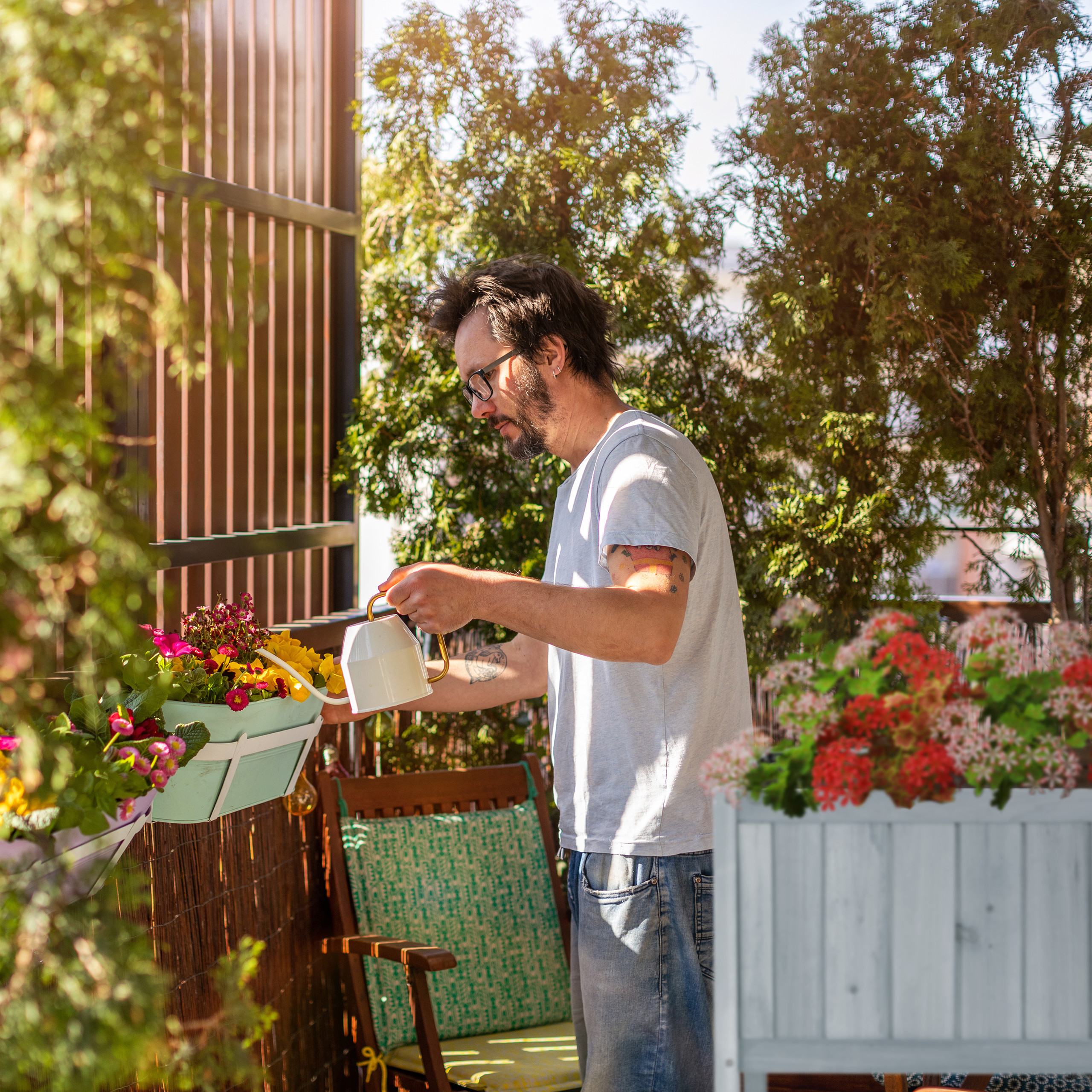Ein Mann mit Brille gießt sonnenbeschienene Blumen auf einem Balkon voller Pflanzen.