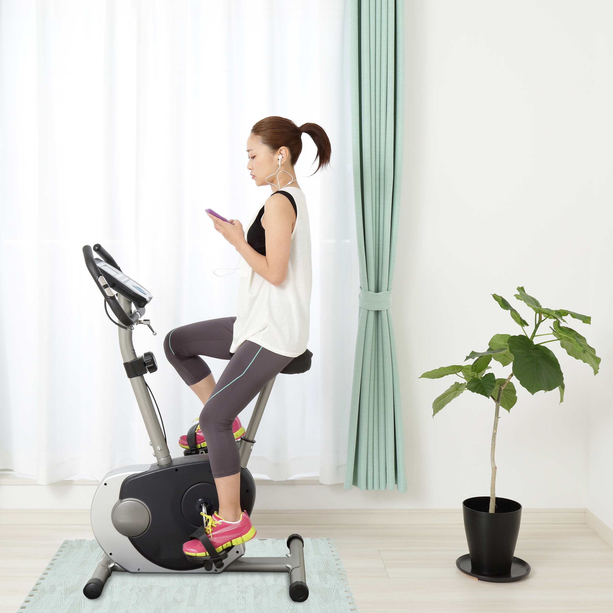Eine Frau nutzt ihr Smartphone und Kopfhörer, während sie auf einem Heimtrainer in einem hellen Raum trainiert.