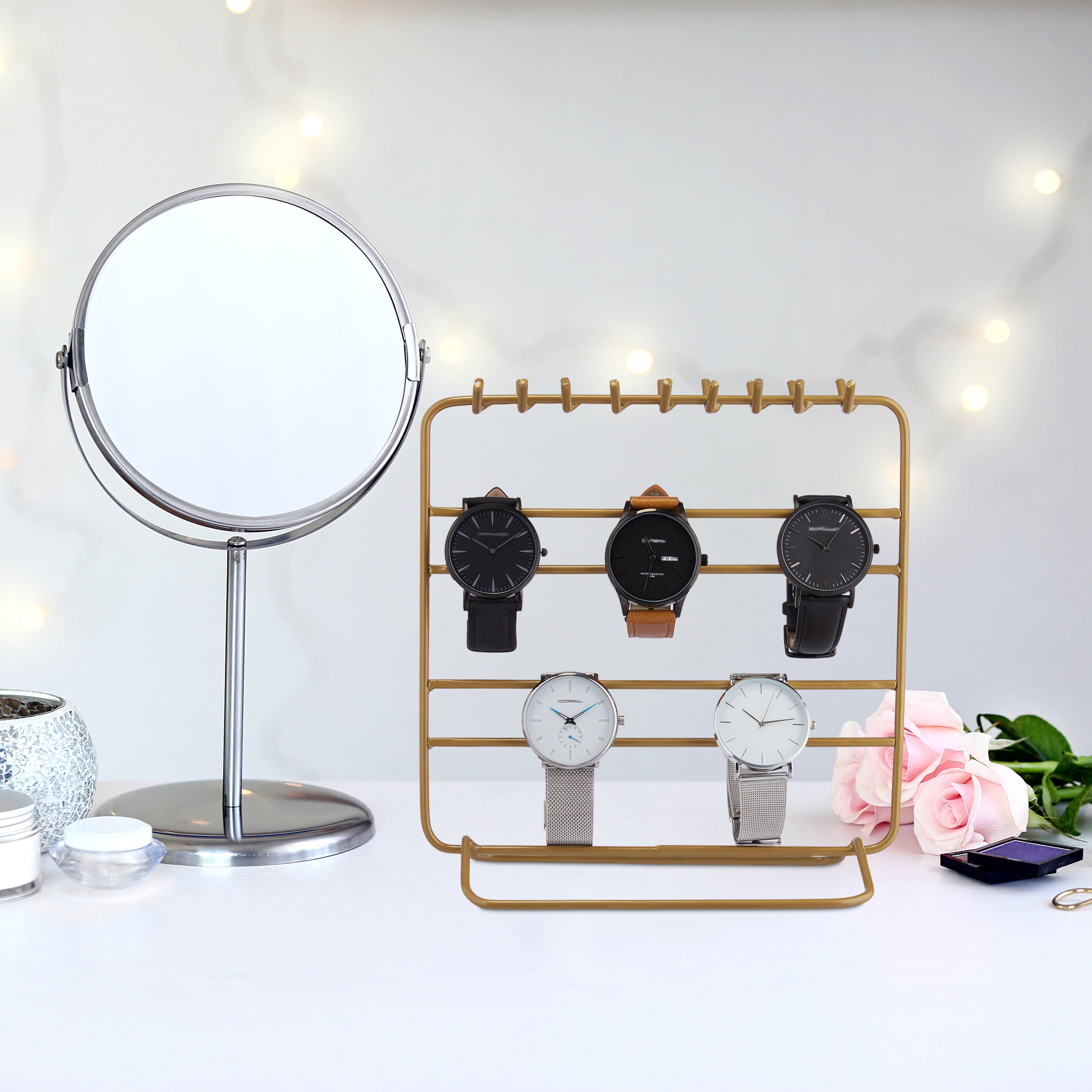 Ein goldener Uhrenständer mit fünf Armbanduhren steht auf einem weißen Tisch neben einem runden Spiegel und rosa Rosen.