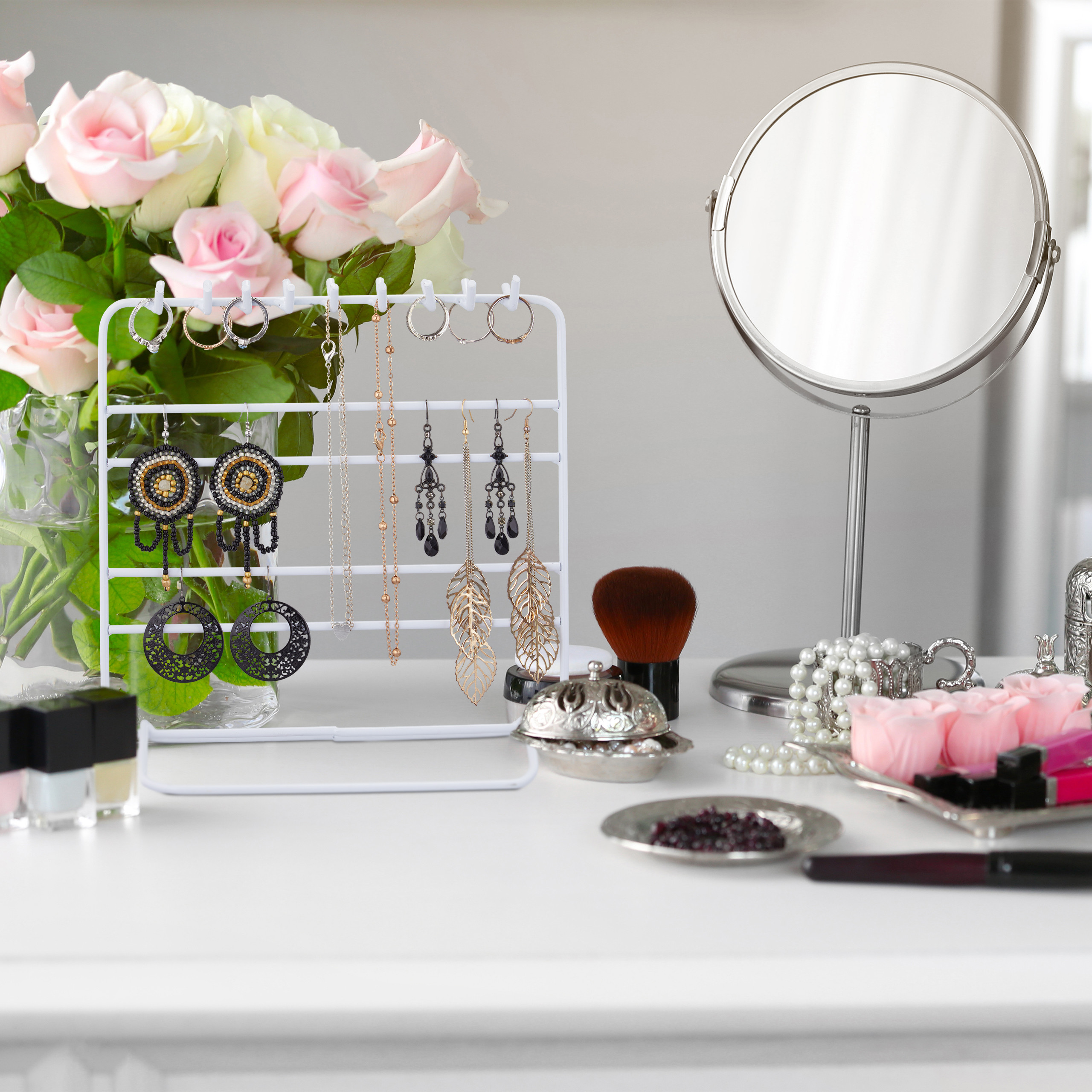 Ein Schmuckständer mit Ohrringen und Ketten steht auf einem Tisch neben Rosen, einem Spiegel und Make-up.
