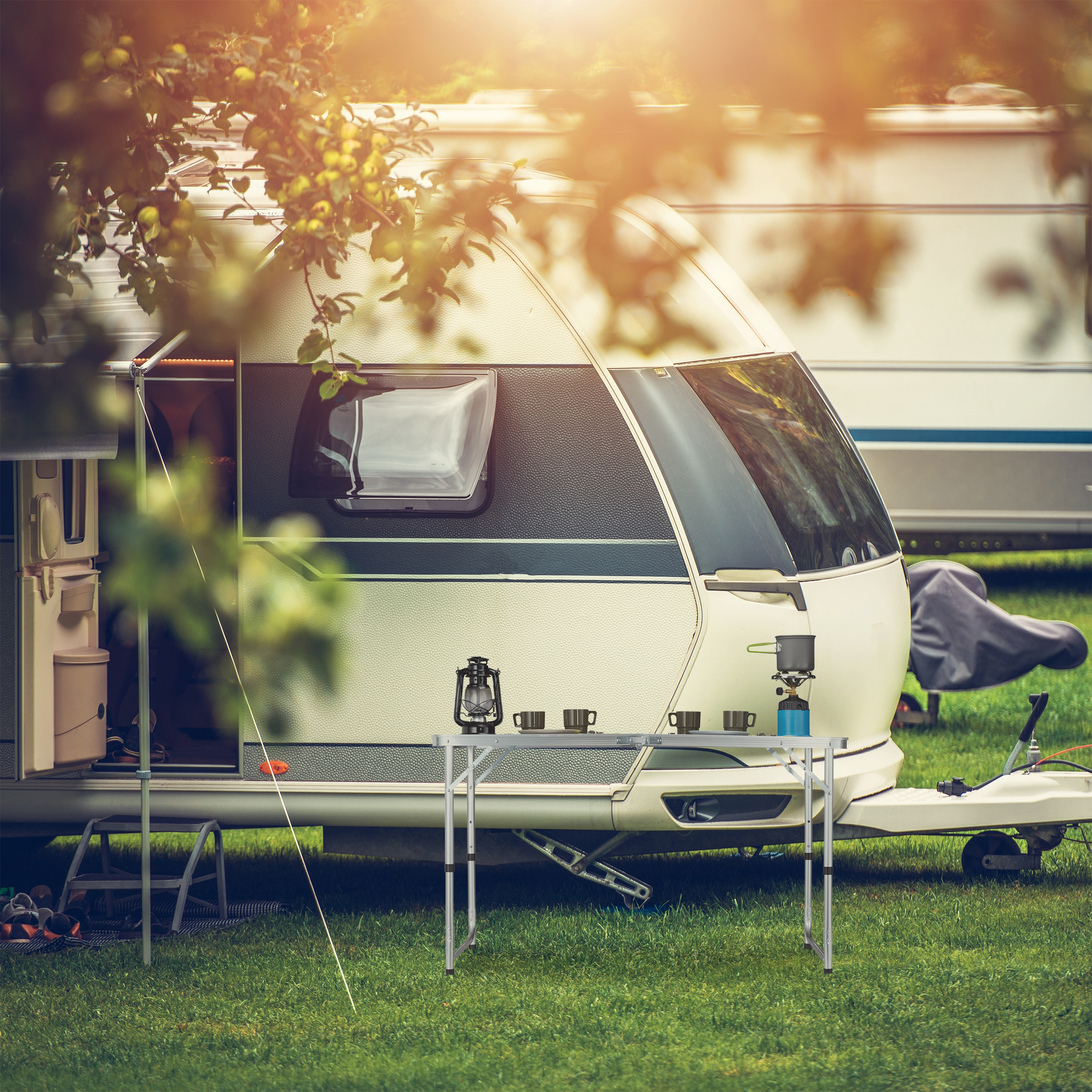 Ein weißer Wohnwagen mit Campingtisch, Tassen und Kocher auf grünem Gras im Sonnenlicht.