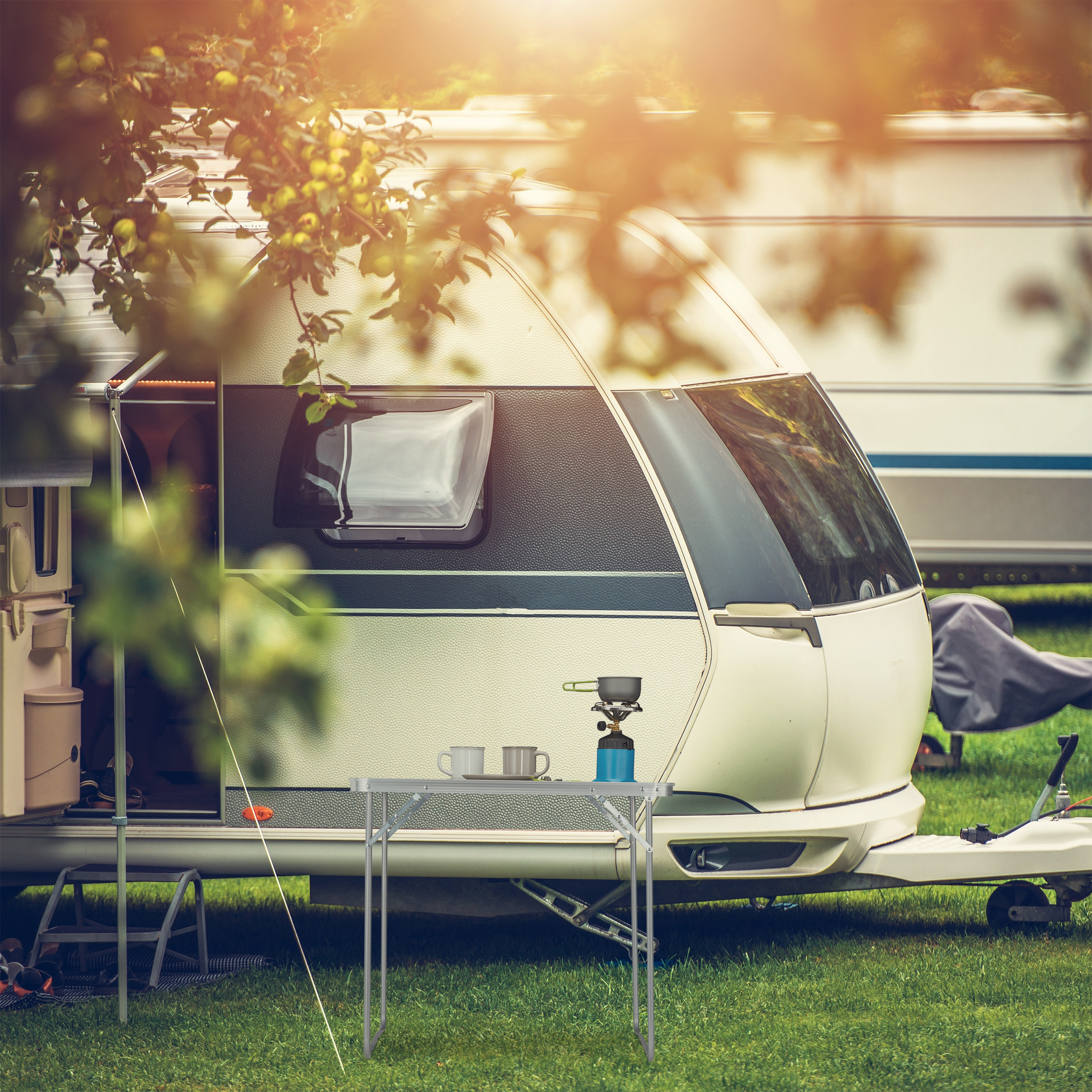 Ein weißer Wohnwagen auf grünem Gras mit einem kleinen Tisch, Campingkocher und Tassen unter einem sonnigen Baum.