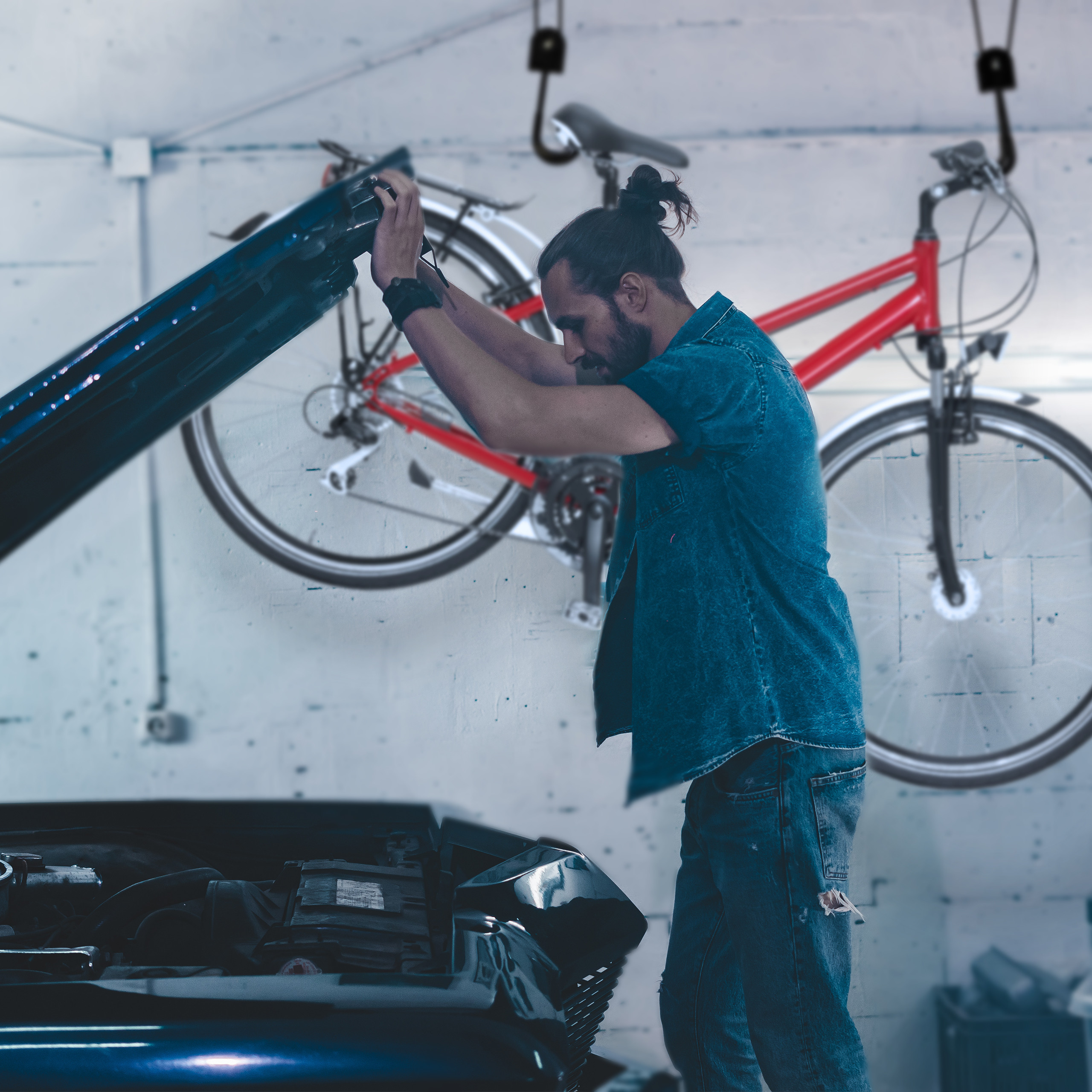 Ein Mann repariert ein Auto in einer Garage, an der Decke hängt ein rotes Fahrrad.