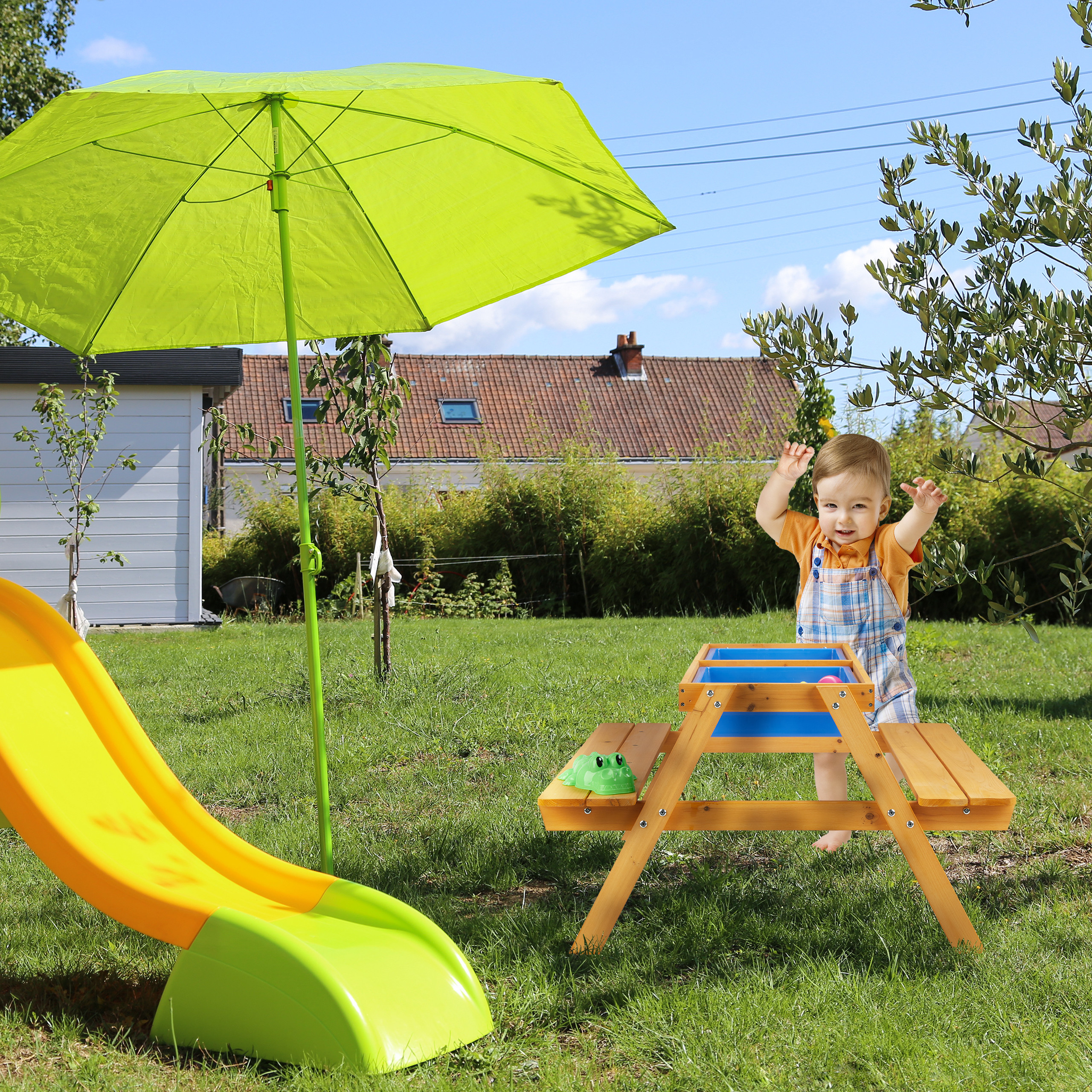 Ein Kleinkind steht neben einem Holztisch mit grünem Schirm und Rutsche auf Rasen im sonnigen Garten.