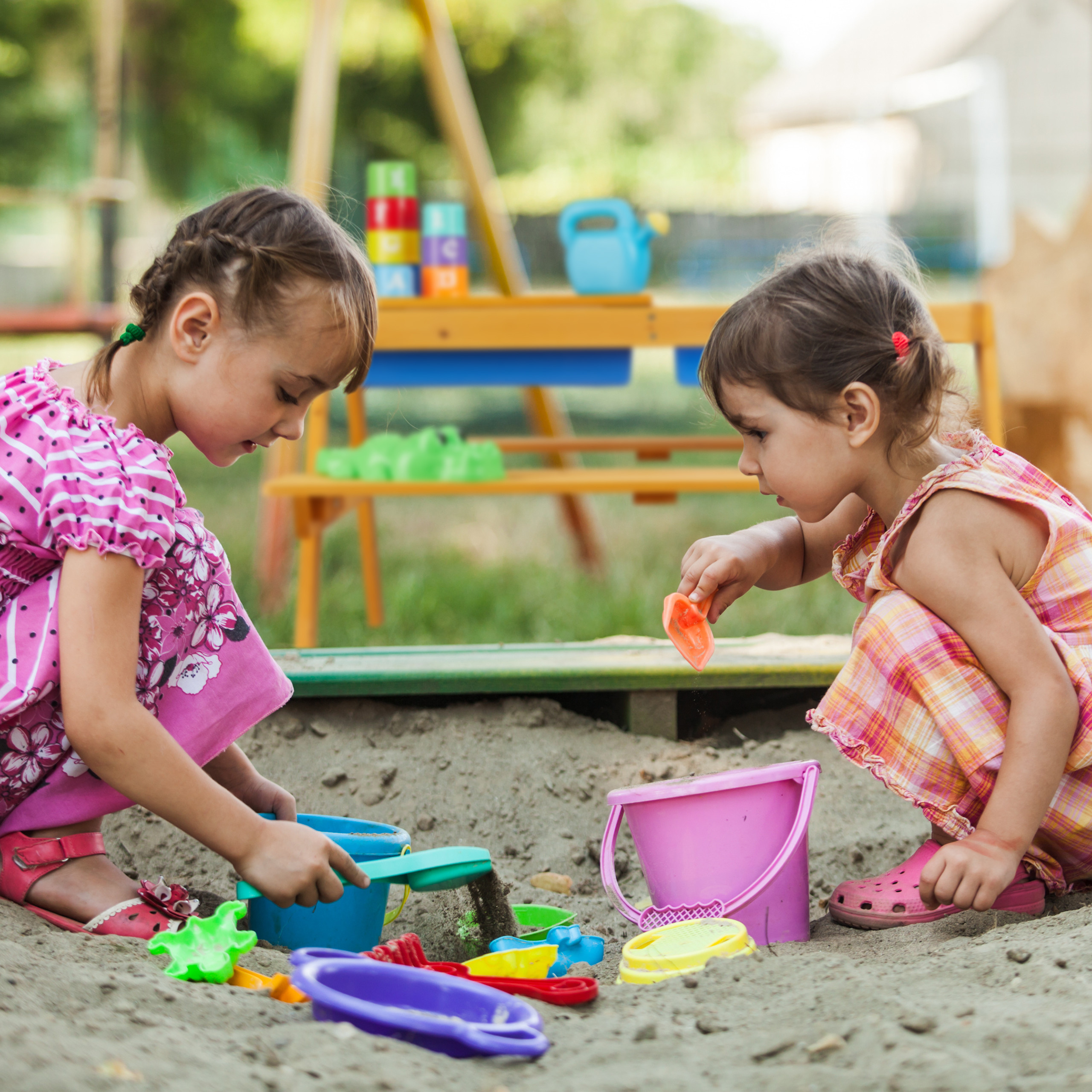 Zwei kleine Mädchen spielen glücklich mit bunten Eimern und Schaufeln im Sandkasten eines Spielplatzes.