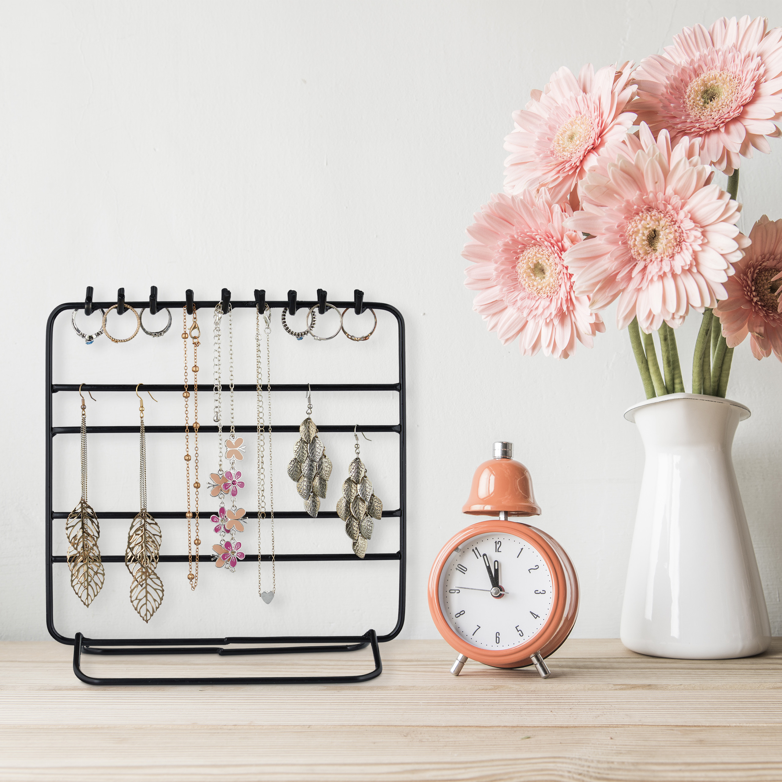 Ein Schmuckständer mit Ohrringen und Ketten steht auf einem Tisch neben einem Wecker und einer Vase mit rosa Blumen.