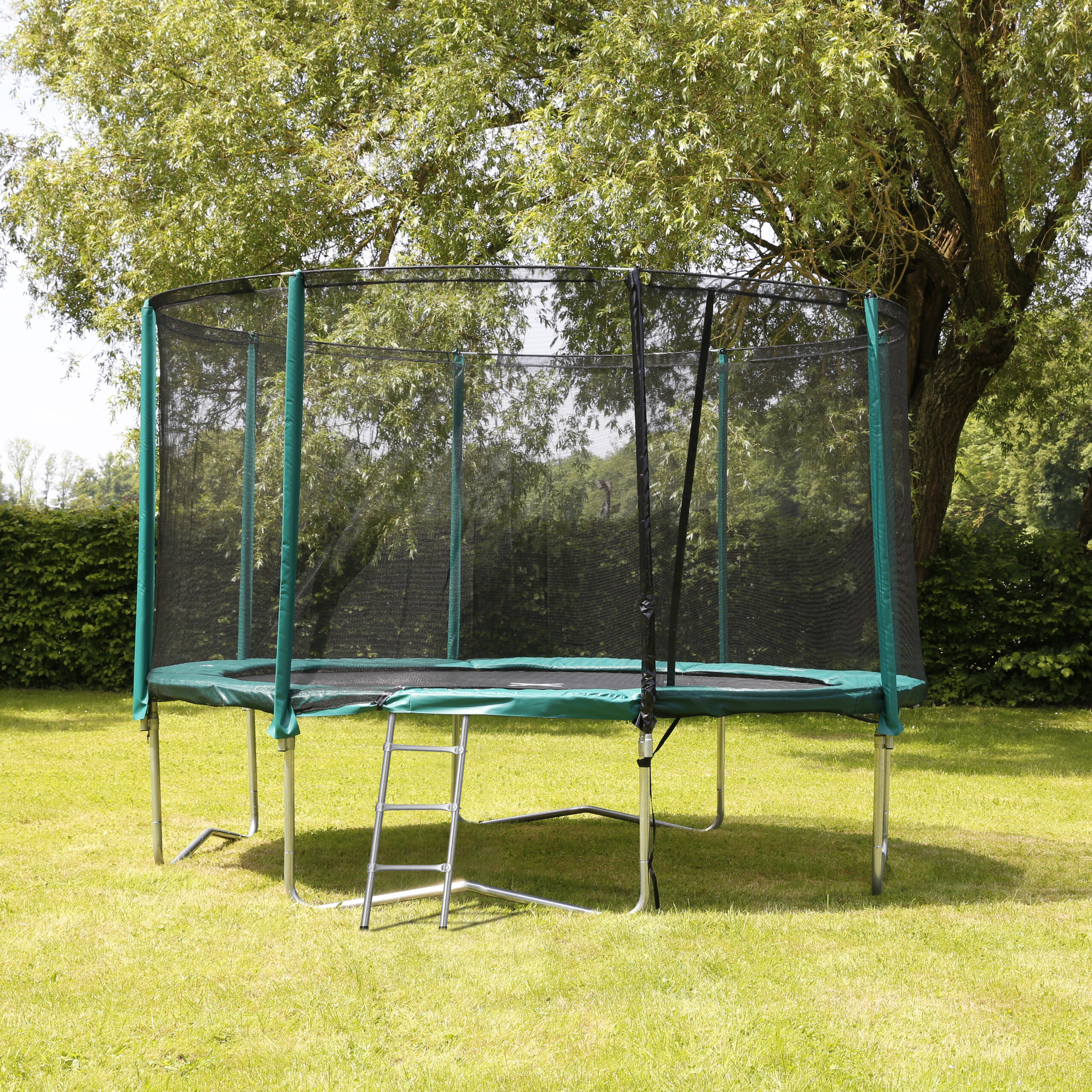 Ein großes grünes Trampolin mit Sicherheitsnetz steht auf einer sonnigen Wiese vor Bäumen.