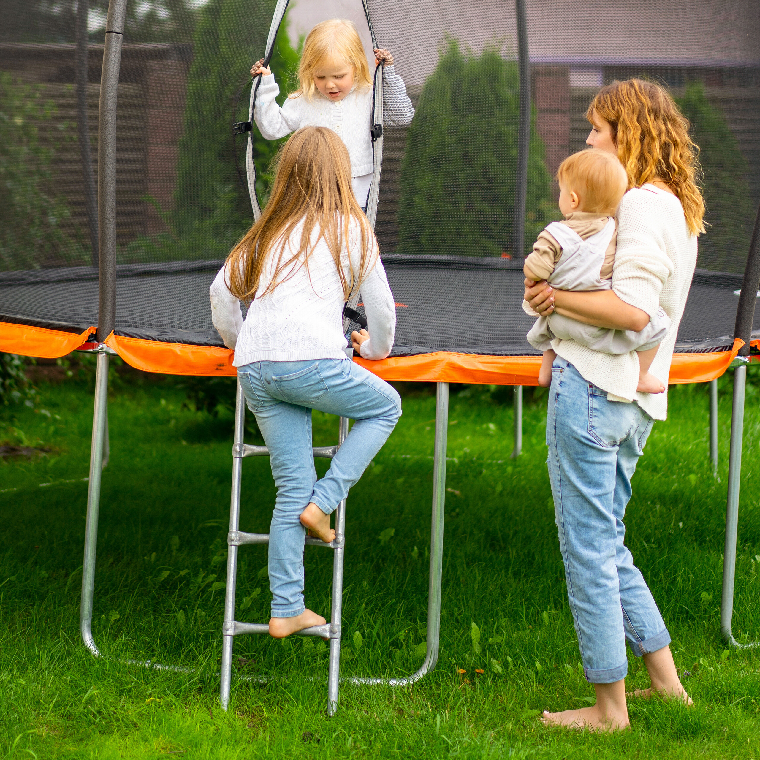 Mutter hält Baby im Arm, während ältere Kinder in Trampolin klettern.