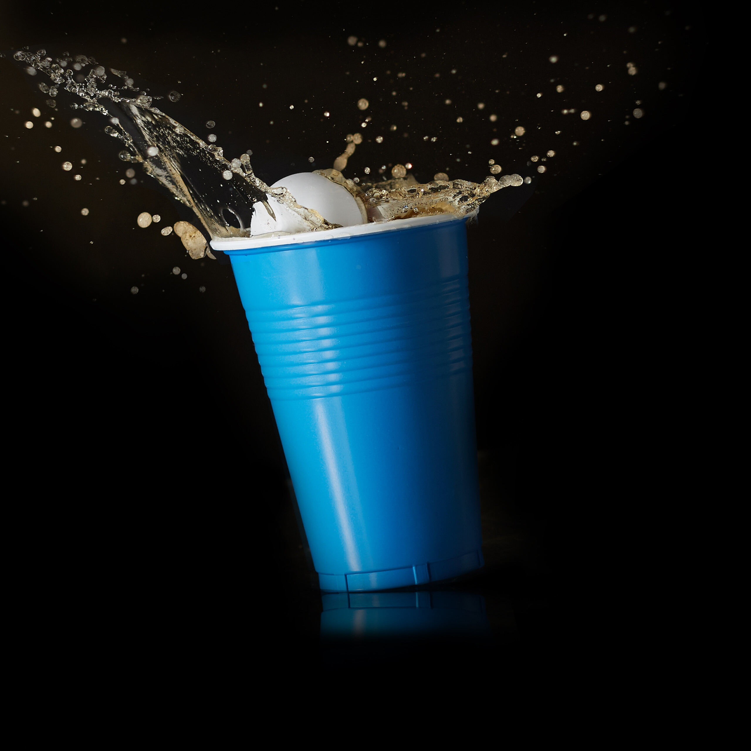 Ein weißer Ball landet in einem blauen Becher und spritzt Flüssigkeit gegen einen schwarzen Hintergrund.