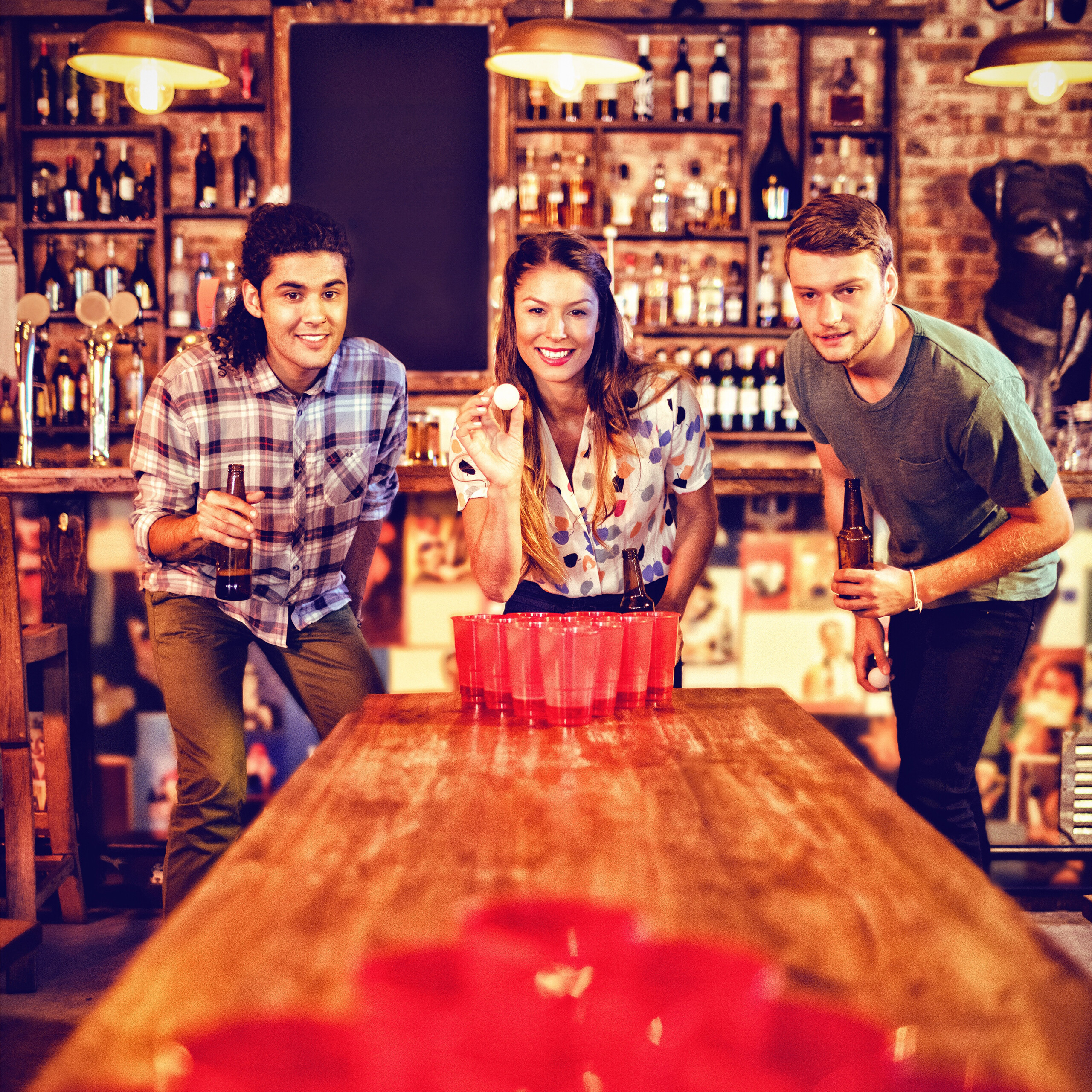 Drei Freunde spielen Bierpong in einer Kneipe, halten Getränke und lächeln an einem Holztisch mit roten Bechern.