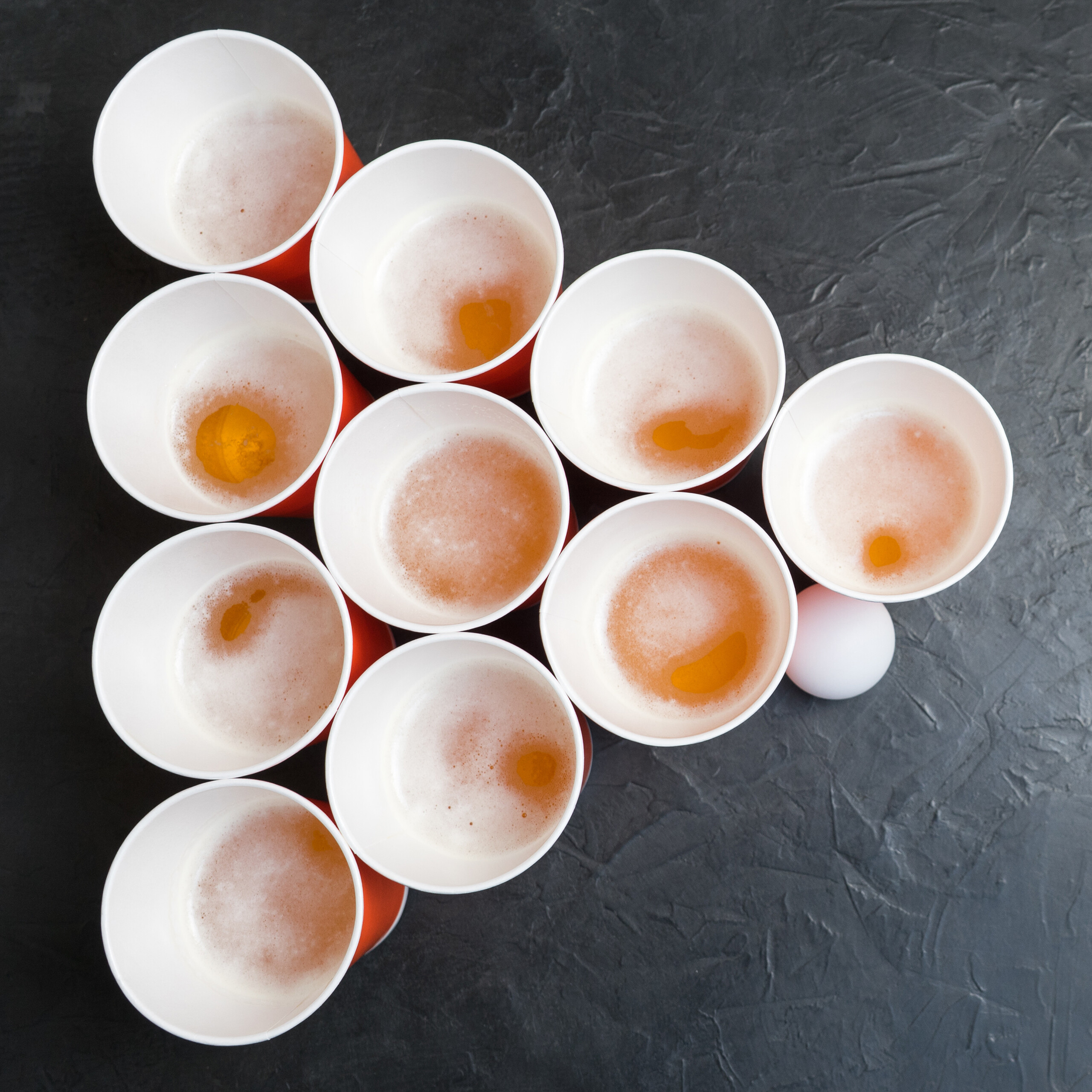 Zehn rote Bierpong-Becher in Dreiecksform auf dunkler Oberfläche, daneben ein weißer Ball.