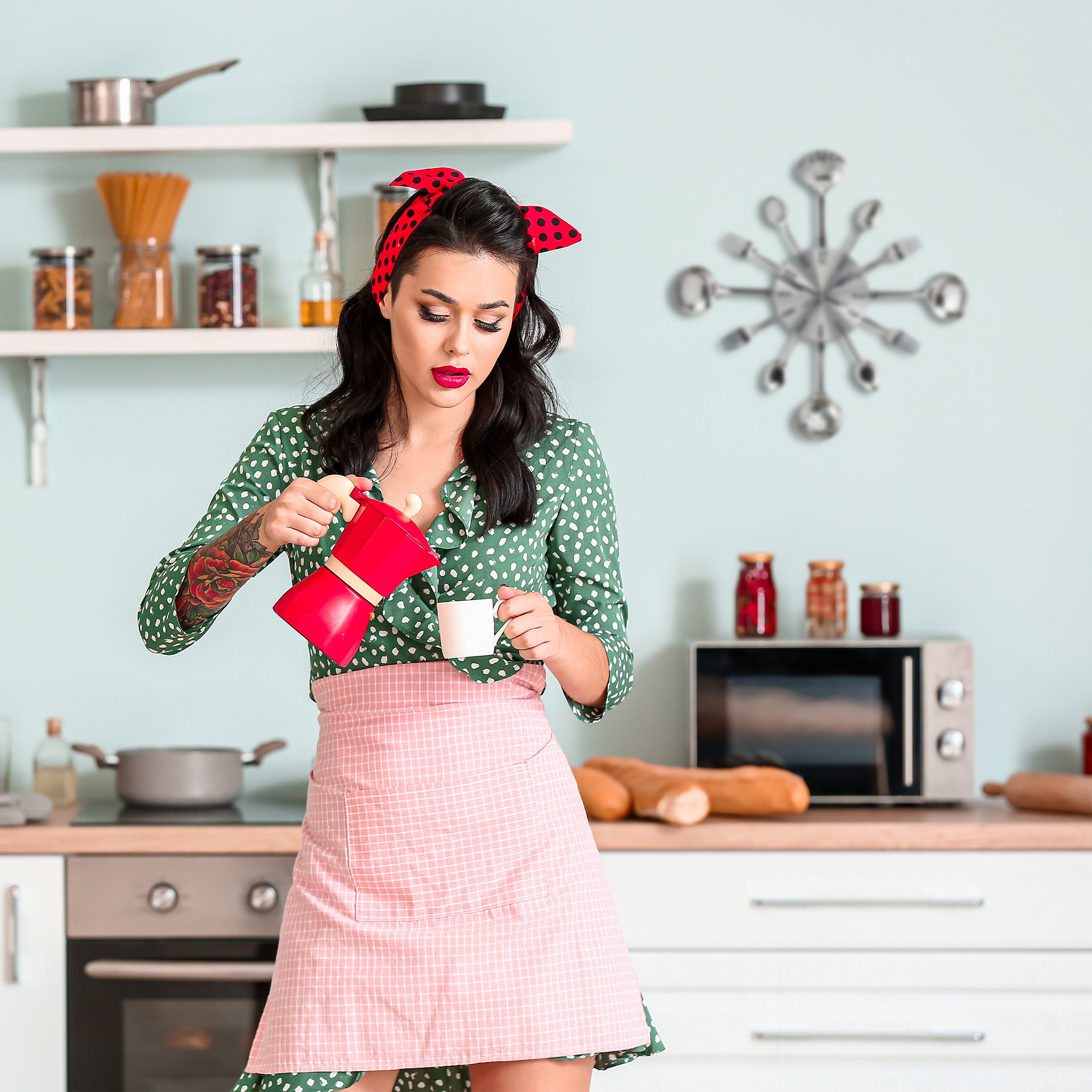 Eine Frau im Pin-up-Stil gießt Kaffee aus einer roten Kanne in eine weiße Tasse in einer hellen Küche.