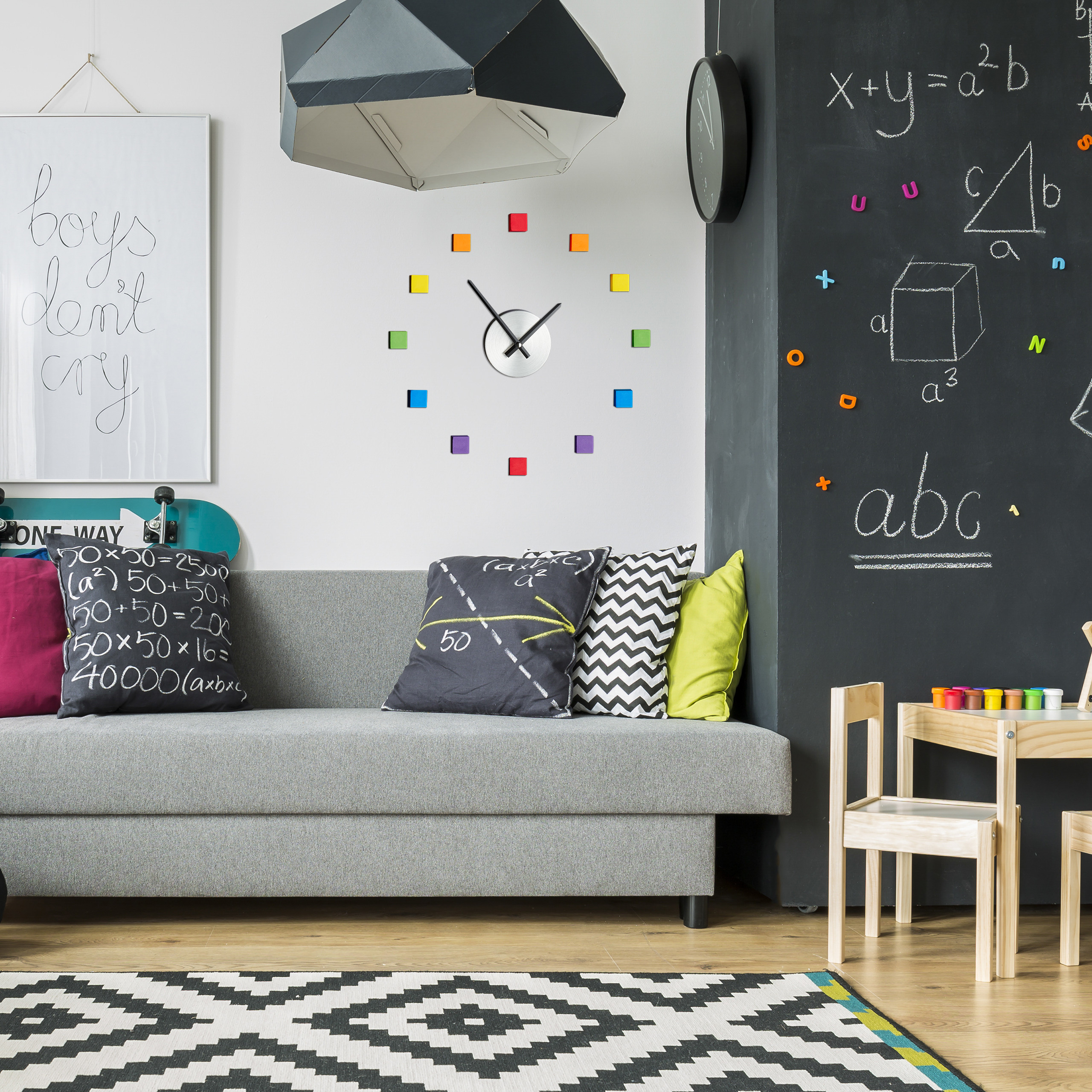 Wohnzimmer mit grauem Sofa, bunten Kissen, farbiger Wanduhr, Wandtafel und Kindermöbeln.