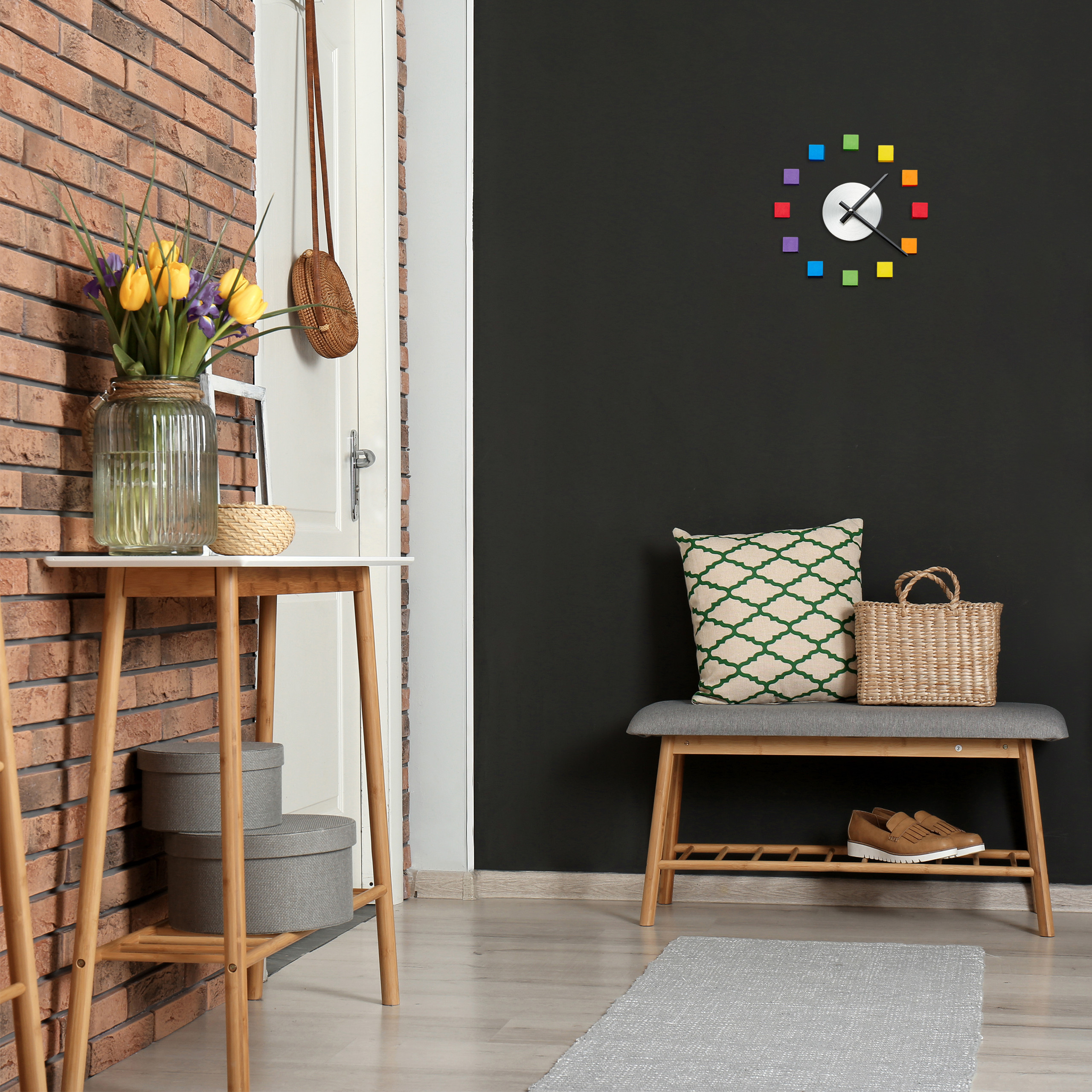 Flur mit Backsteinwand, Konsole mit gelben Blumen, Holzbank mit Kissen und bunter Uhr an schwarzer Wand.