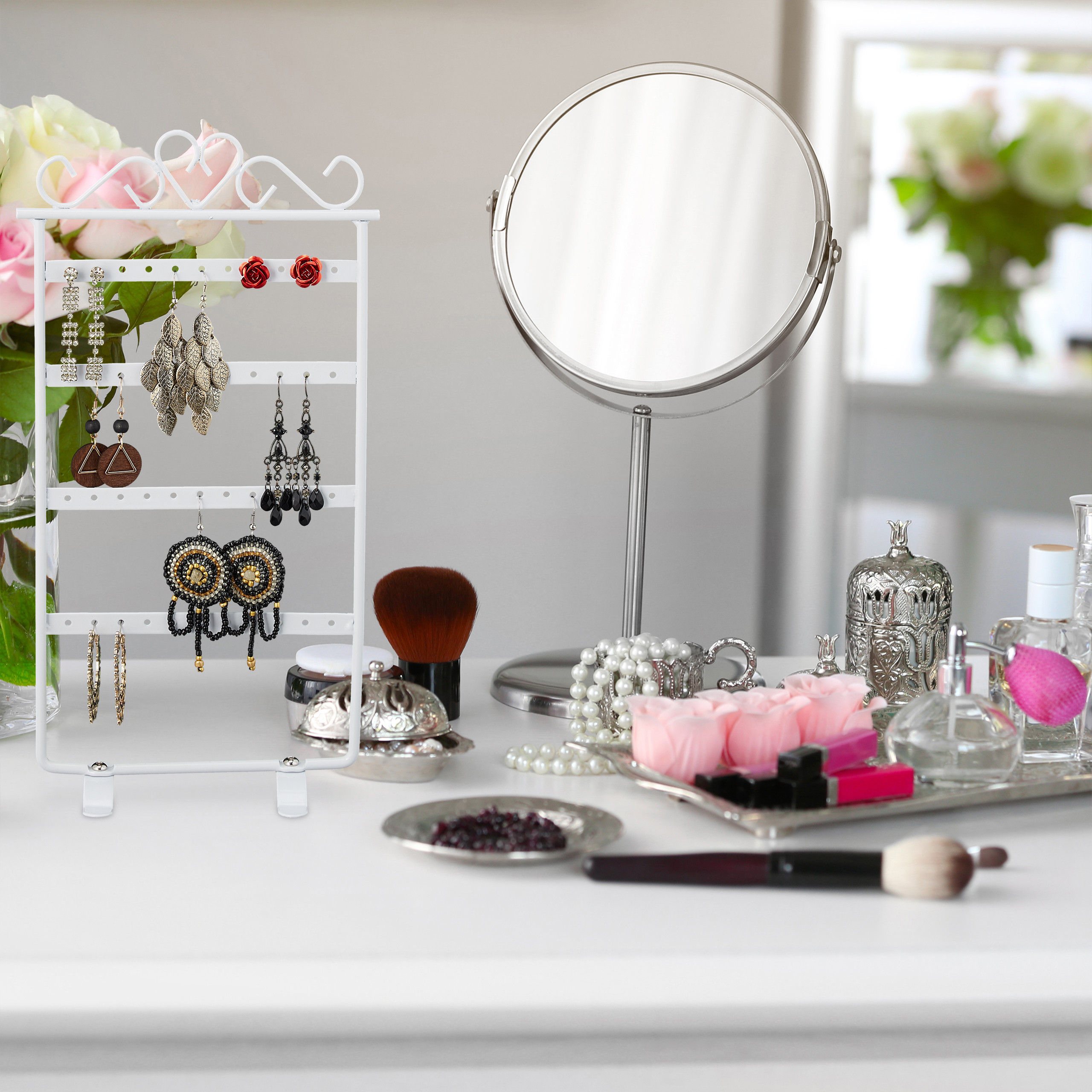 Ein Schminktisch mit Ohrringen, einem runden Spiegel, Make-up-Pinseln und Parfümflaschen vor Blumen.