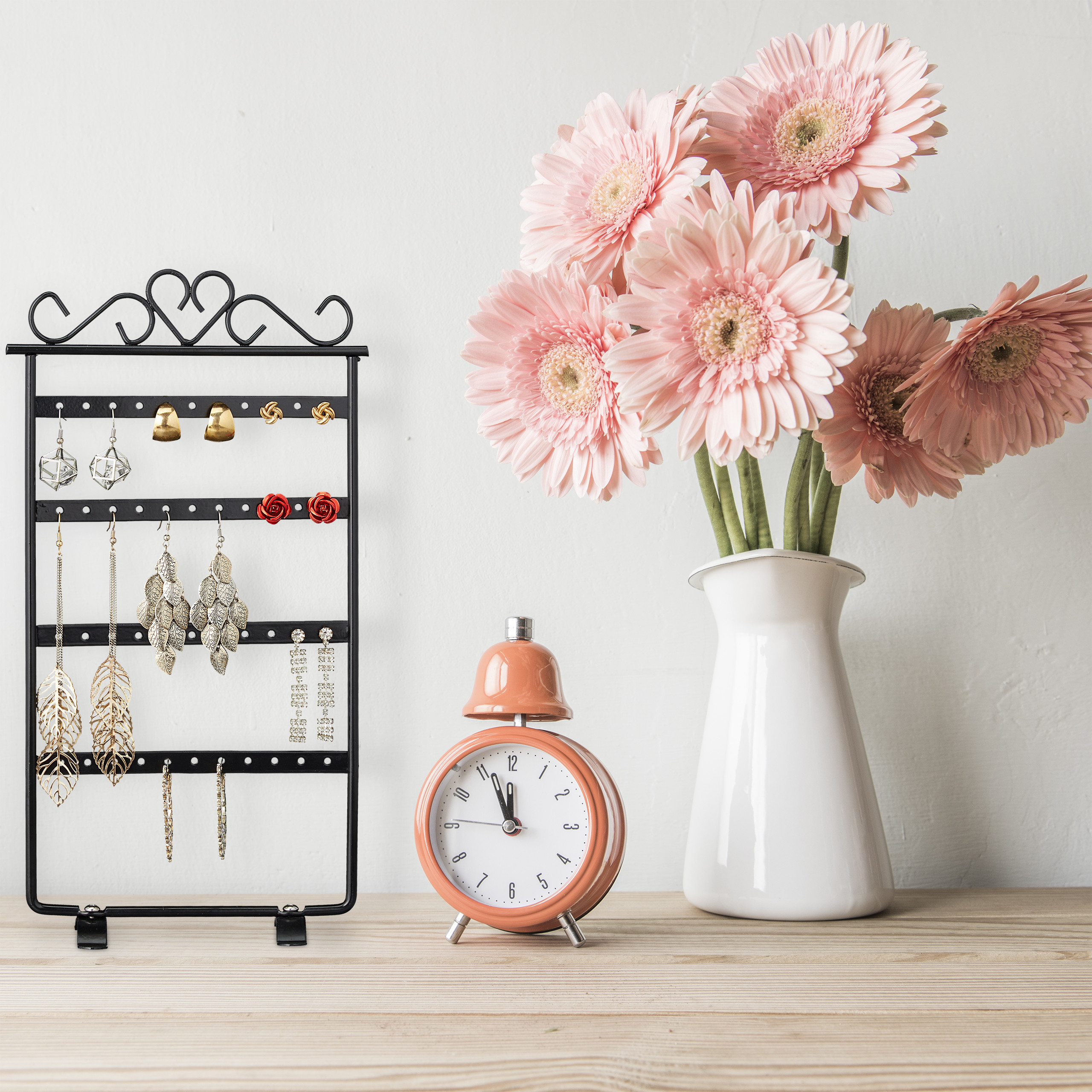 Ein schwarzer Schmuckständer, ein Wecker und rosa Gerbera in einer Vase auf einem Holztisch.