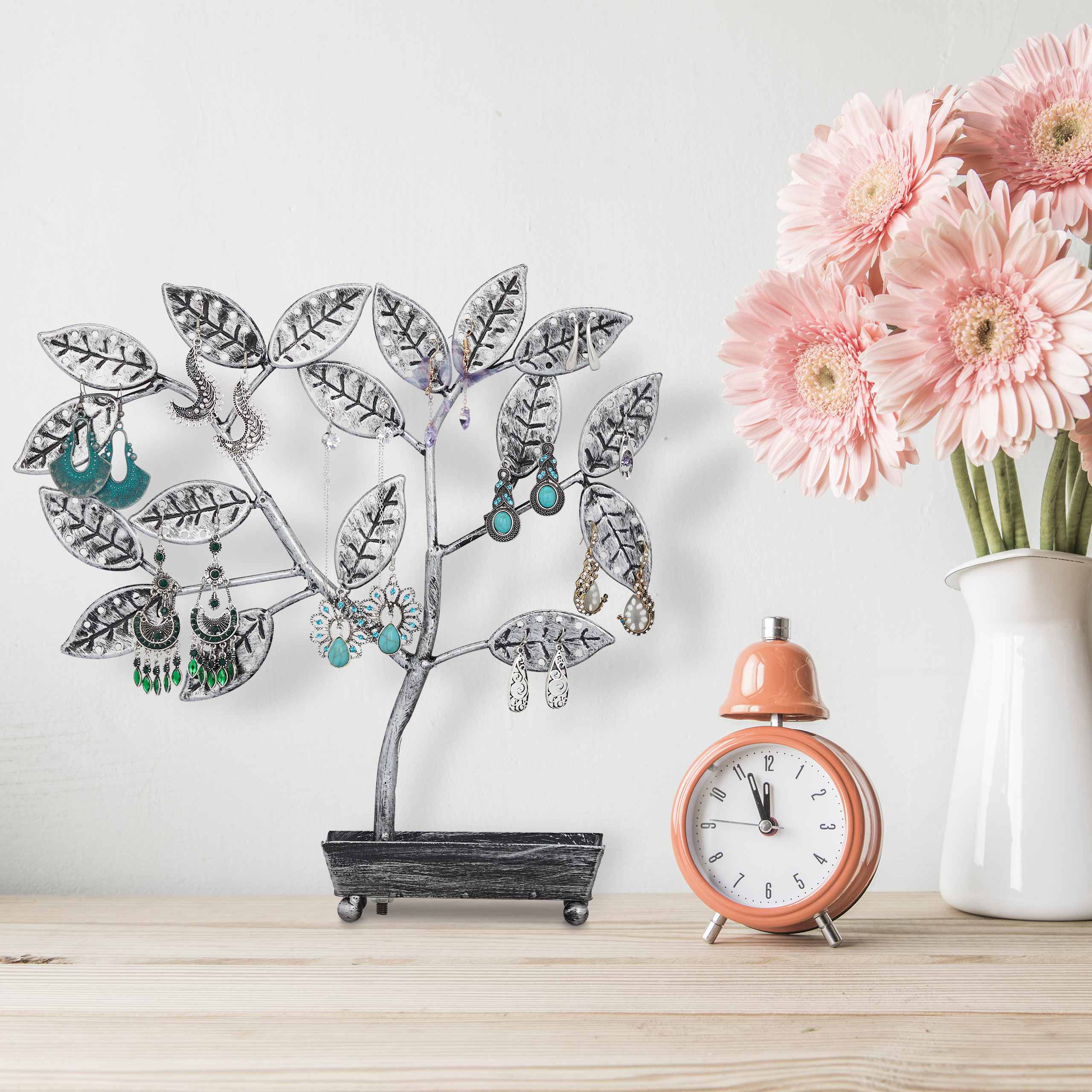 Ein Schmuckhalter in Baumform mit Ohrringen, ein pinker Wecker und rosafarbene Blumen in einer weißen Vase auf einem Tisch.